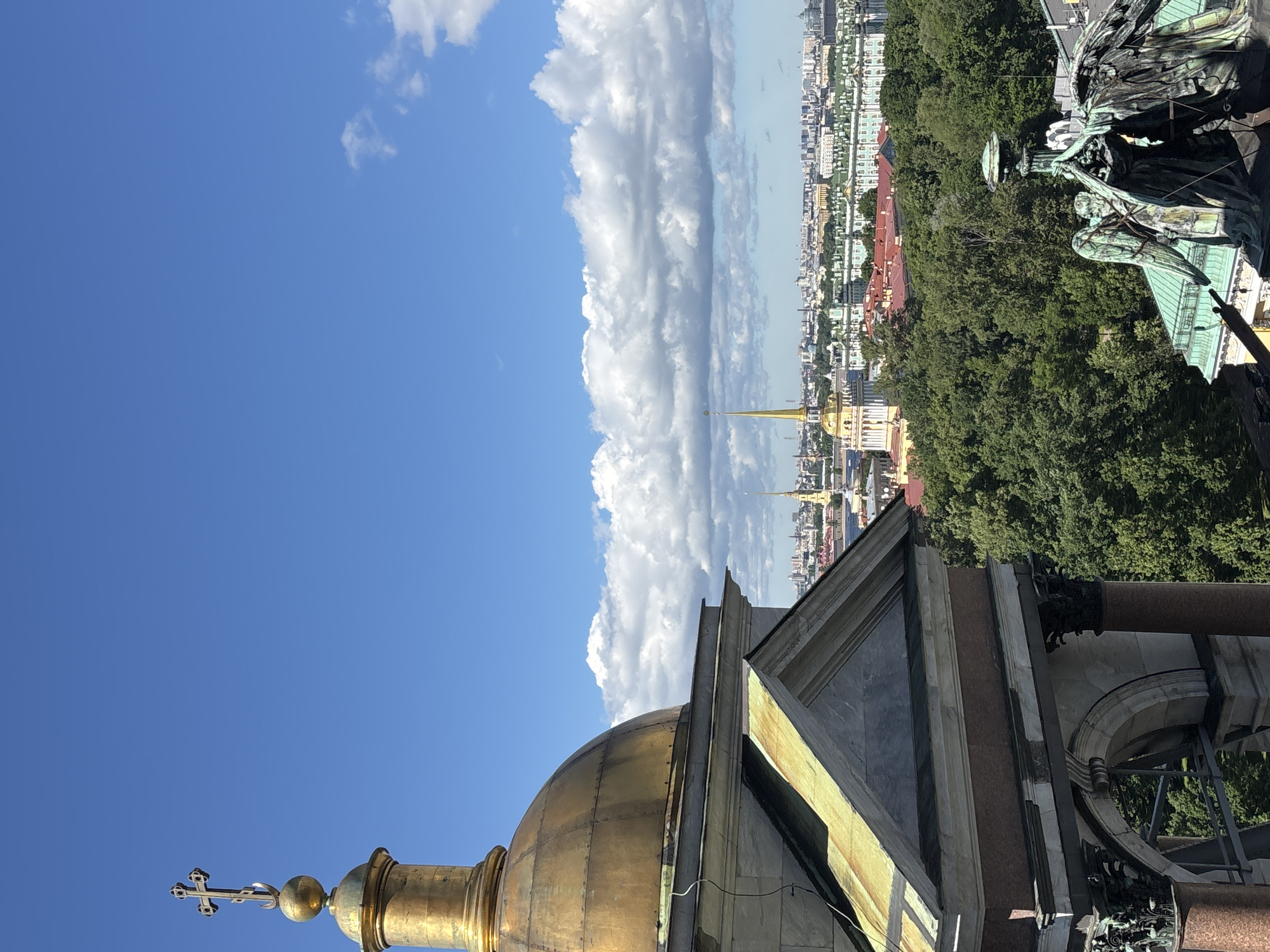 This media file is a vertical, wide-angle photograph taken from a high vantage point, offering a panoramic view of a cityscape under a bright, partly cloudy sky. The foreground features the ornate golden dome of a building, topped with a cross. Adjacent to the dome, a section of stone architecture with triangular pediments and decorative elements is visible.
The middle ground reveals a densely built urban landscape with a mix of building styles and colors, suggesting an older city. Tall, slender spires, some golden, punctuate the skyline. Lush green trees are interspersed throughout the urban fabric, creating a contrast between nature and the built environment.
In the upper right portion of the image, a large, detailed sculpture of winged figures, possibly angels, is visible, contributing to the sense of grandeur and possibly historical significance. The sculpture appears to be made of a dark, weathered metal.
The sky is a vibrant blue, with large, fluffy white clouds dominating the left side of the frame and stretching horizontally across the scene. The lighting suggests it is daytime, likely late morning or early afternoon, with the sun casting distinct shadows.
The overall impression is one of an expansive, historic European city, viewed from a significant height, perhaps from the top of a cathedral or tower. The weather appears pleasant, with clear skies and some clouds adding visual interest. There is no visible text in the image.