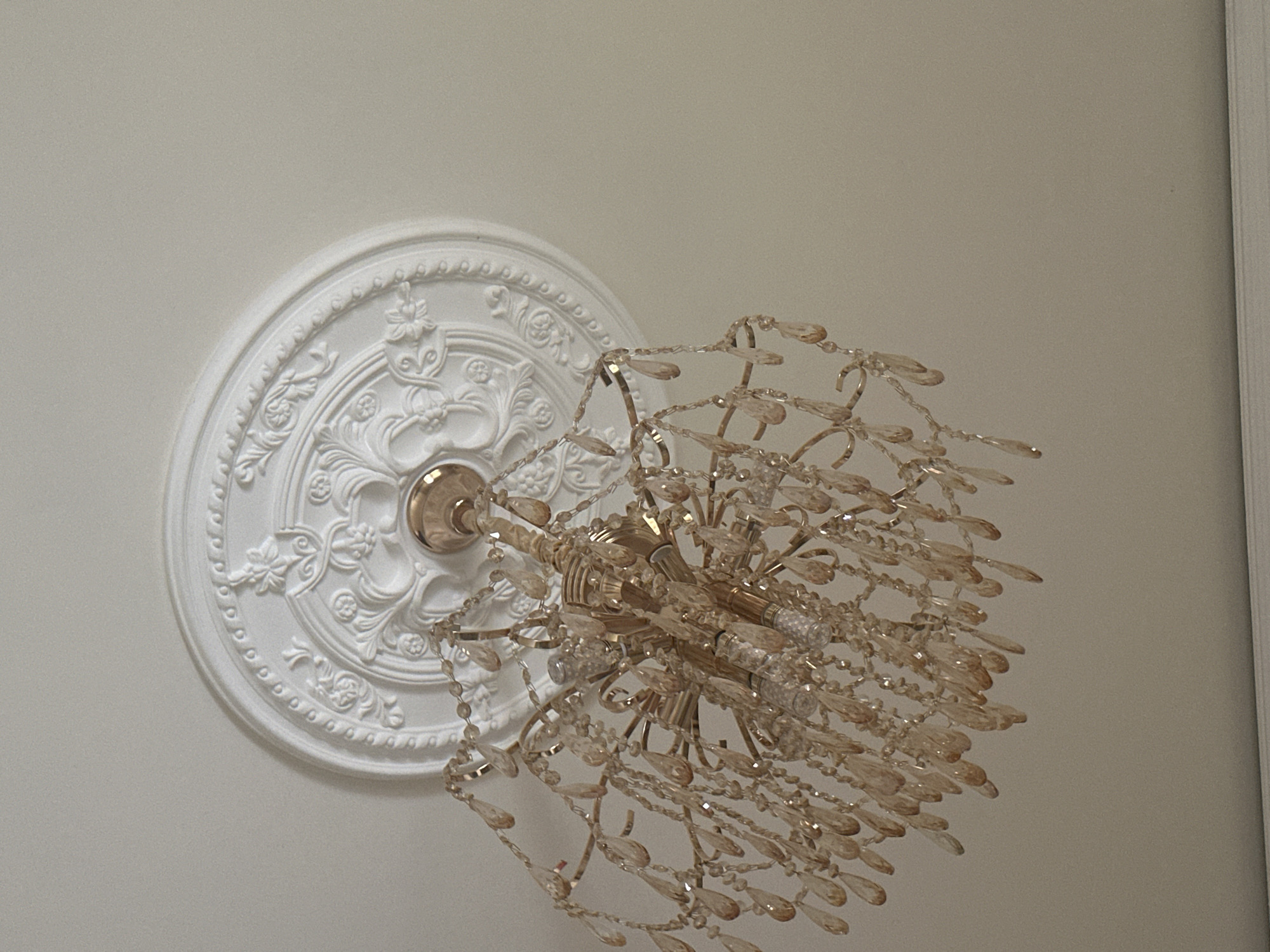 An ornate, gold-colored chandelier hangs from a white ceiling medallion in an indoor setting. The chandelier features multiple tiers of teardrop-shaped amber crystals and has visible light bulbs. The ceiling medallion is round with intricate relief patterns, including floral motifs and beaded edges. The background is a plain, off-white ceiling. The image is a close-up shot looking upwards at the chandelier and ceiling fixture.