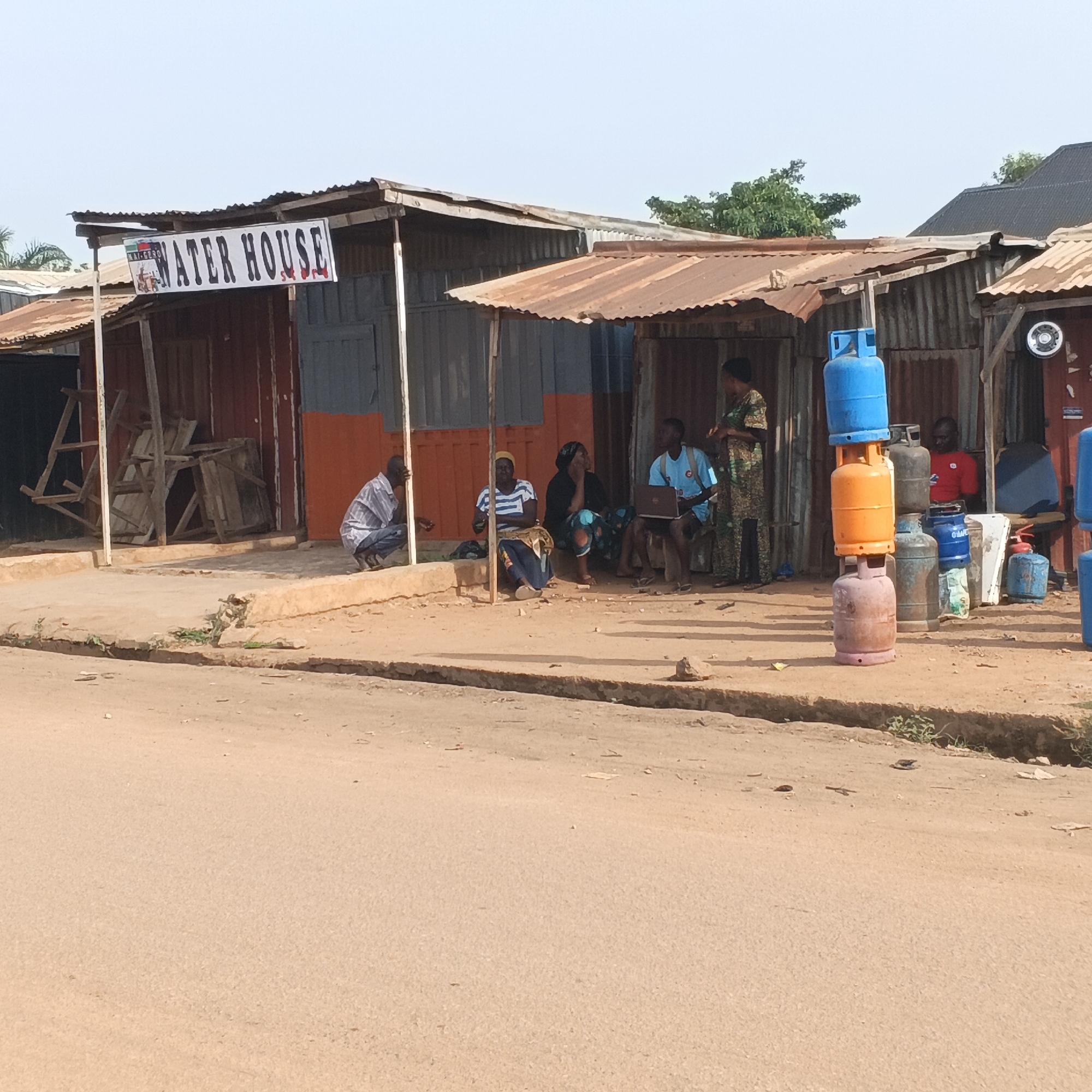 This image depicts a street scene in Rido, Nigeria. A row of makeshift structures with corrugated metal roofs lines a dirt road. A sign above one of the structures reads "WATER HOUSE." In front of these structures, several people are gathered.
On the left side, a man in a striped shirt and jeans squats near a white pole. Next to him, three people are seated: a woman in a striped shirt and blue skirt, a woman in a black headscarf and patterned skirt, and a young man in a blue t-shirt who is using a laptop. Further to the right, a woman in a patterned dress stands, possibly interacting with the man using the laptop.
In the foreground, stacked gas cylinders are visible, with a blue one at the top, an orange one in the middle, and a reddish-brown one at the bottom. Other gas cylinders and various objects, including a red fire extinguisher, are also present near the structures. The environment appears dusty and sun-drenched, with sparse vegetation growing along the roadside. The overall impression is that of a local marketplace or service area.