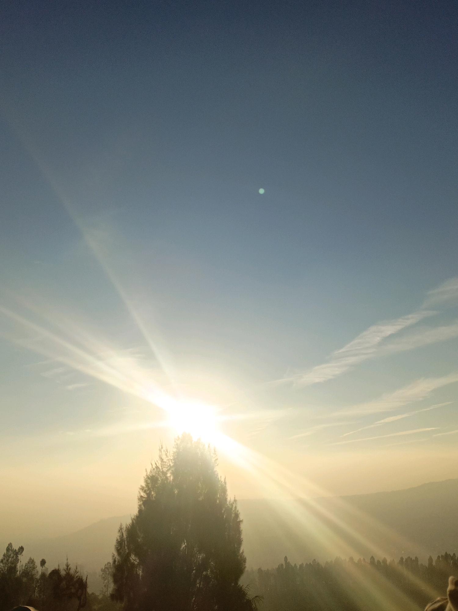 A bright, sunlit sky dominates the upper half of the image, with rays of light bursting from the lower right. A faint, greenish-blue orb is visible in the upper center of the sky. The lower half of the image shows a silhouetted landscape of trees and rolling hills, bathed in the warm glow of the sun. The scene appears to be a natural outdoor setting, possibly a viewpoint overlooking a valley or countryside. There are no people or specific city landmarks visible in the image.