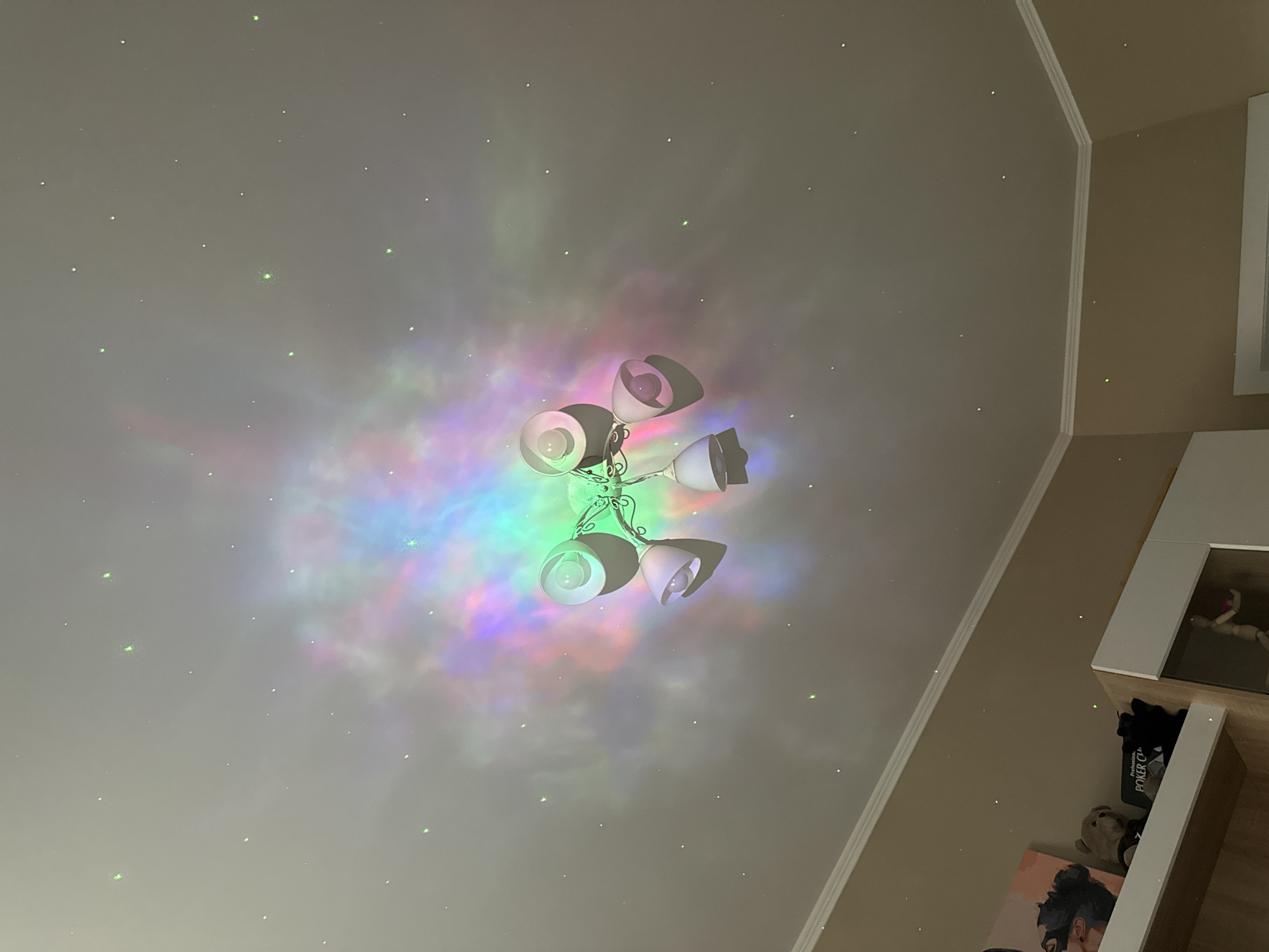 The image shows a ceiling with a light fixture. The light fixture has multiple bulbs and ornate metalwork. The ceiling is white and has a multicolored light projection with star-like points. The angle suggests the image was taken looking upwards. In the lower right corner, there is a glimpse of a shelf with items on it, including a book or game titled "Poker CH". The environment appears to be a residential interior. The location is unknown.