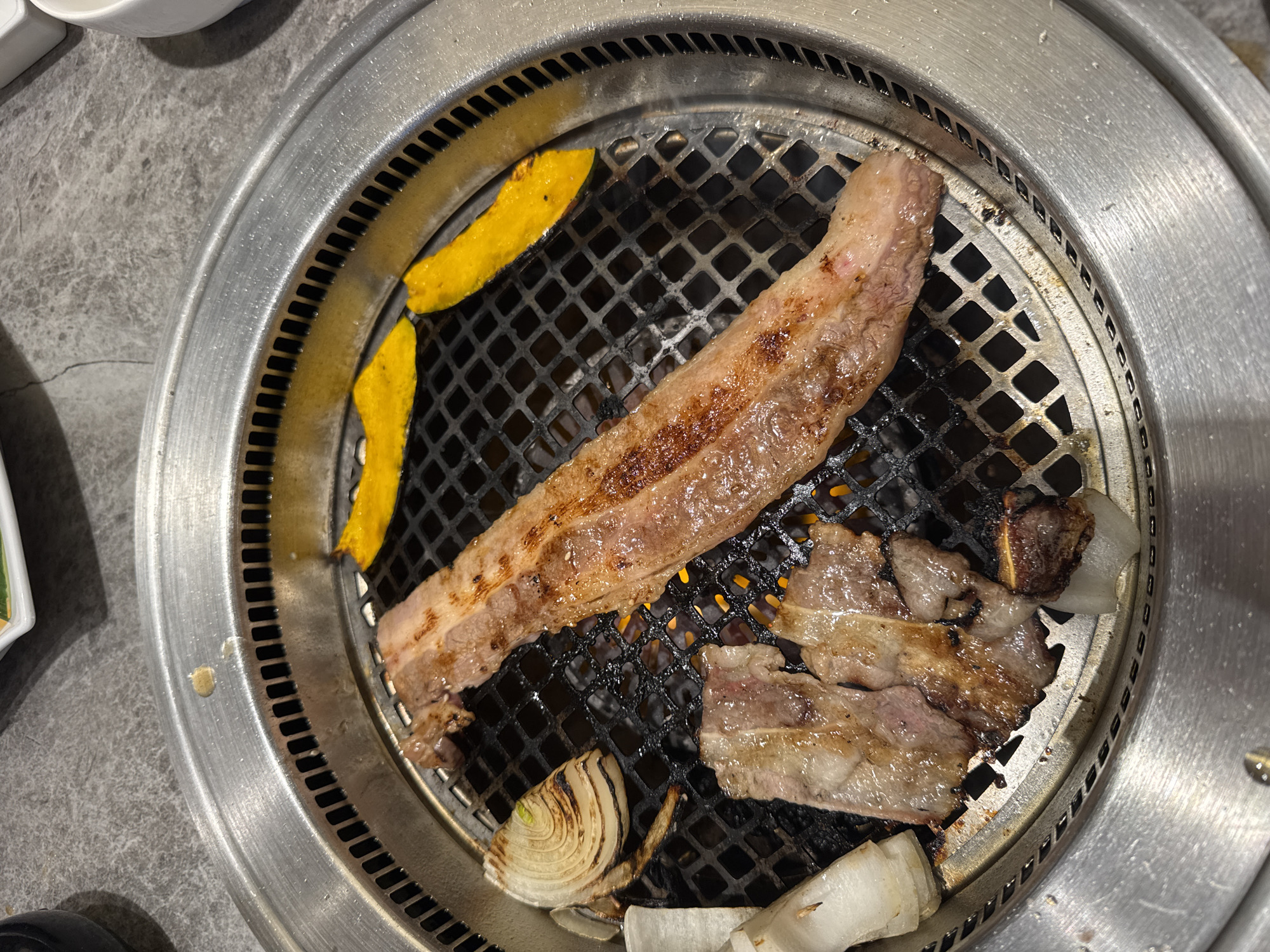 An overhead shot captures a round, metallic tabletop grill filled with food being cooked. The grill features a dark, square-patterned grate, beneath which an orange glow from the heat source is visible.
Several cuts of meat are arranged on the grill. A long, flat strip of well-seared meat, likely pork belly or beef, extends diagonally across the upper left section of the grate, exhibiting browned edges and some charring. To its lower right, several smaller, thinner slices of meat are also cooking, showing varying degrees of browning and pinker, uncooked areas.
Accompanying the meat are vegetable slices. Two bright yellow, crescent-shaped pieces, resembling pumpkin or squash, are placed on the upper left side of the grill. At the bottom, several slices of white onion are visible, some already softened and slightly charred from the heat.
The circular grill is integrated into a grey, textured tabletop, which shows some visible wear and smudges. The metallic rim of the grill has some grease splatters. The scene depicts an ongoing BBQ or grilling session, commonly found in Korean or Japanese-style restaurants, in what is likely an indoor dining setting in Kuta Selatan, Indonesia. No people or visible text are present in the image.