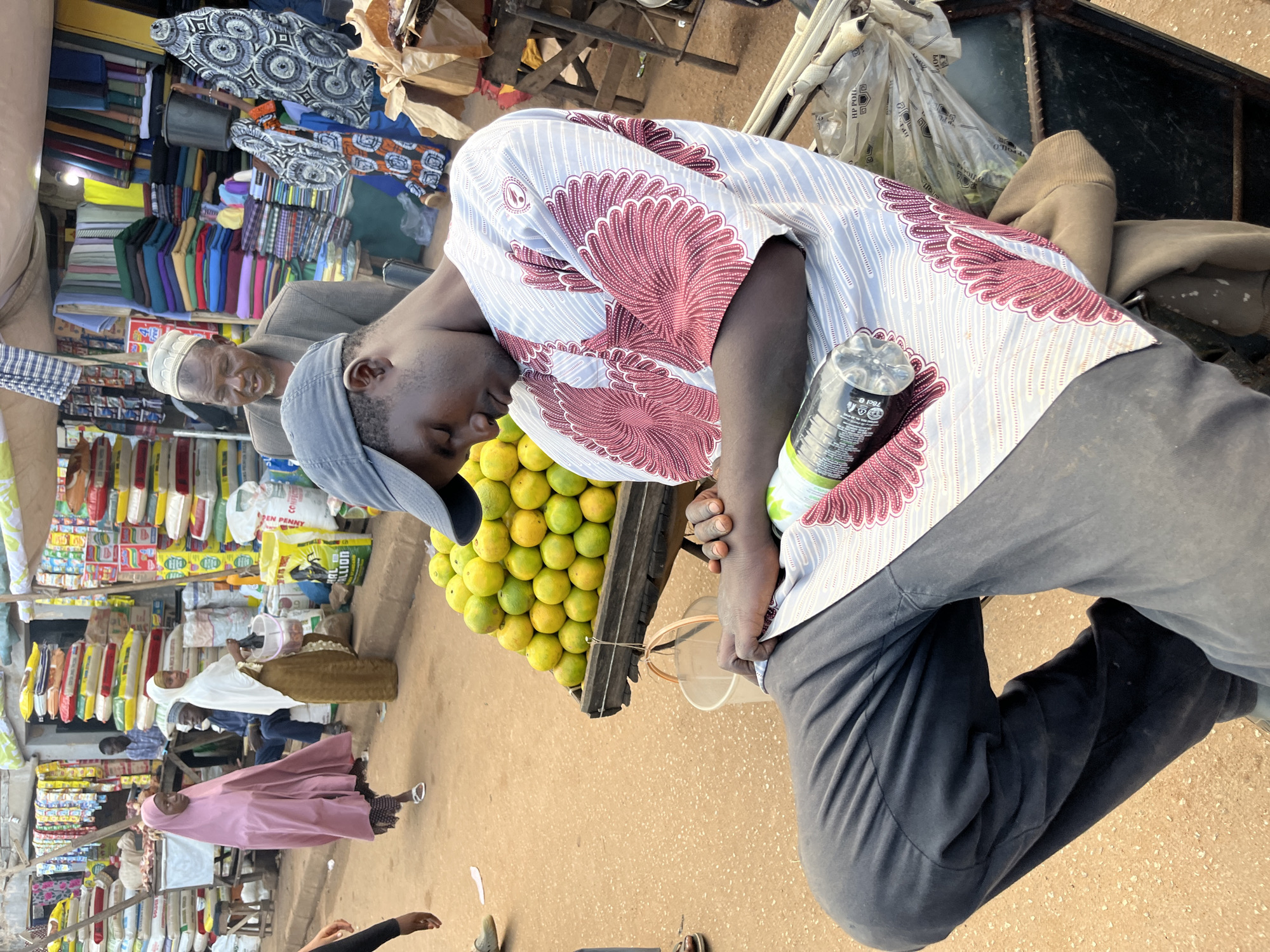 In Bukuru, Nigeria, a man wearing a gray baseball cap and a white and red patterned shirt is asleep, slumped over a cart filled with oranges. He has a black bottle of water lying beside him. In the background, a bustling marketplace is visible, with stalls selling fabrics, grains, and other goods. Several people, including two women wearing headscarves and an elderly man in a cap and jacket, are browsing the merchandise. The ground is dusty, indicating an outdoor setting.