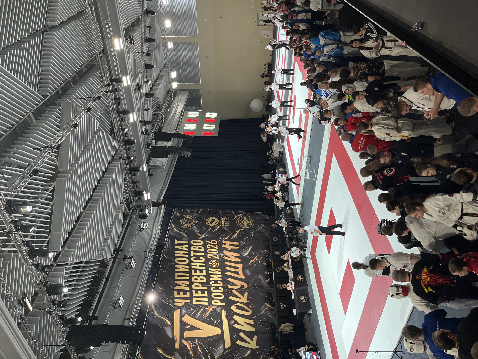 The image captures an indoor martial arts competition, specifically the "Russian Championship Championship 2026 Kyokushin" held in what appears to be Zhavoronki, Russia. A large banner displaying the event's name in Russian dominates the lower portion of the frame. Red and white striped mats are visible, with rows of seated competitors in white and blue uniforms on either side. Several individuals in white shirts and black pants are seen standing on the mats. A scoreboard hangs above the scene. The venue has a complex, modern ceiling structure with metal beams and lighting fixtures.