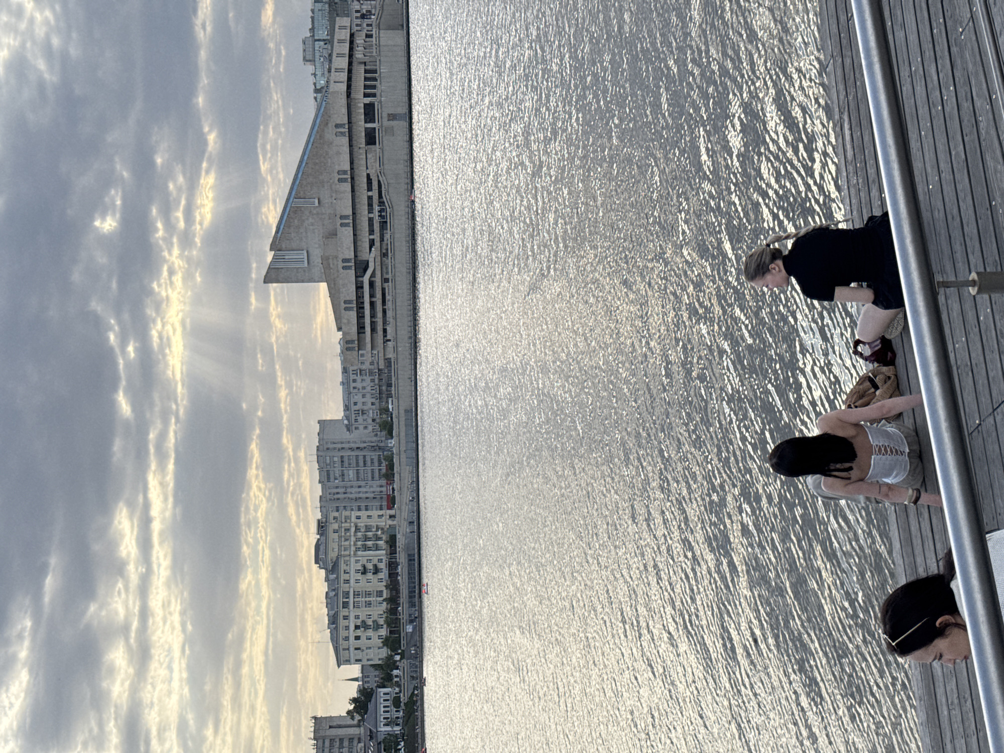 The image captures a scene along the waterfront in Kazan, Russia. Three women are positioned on a wooden boardwalk or pier. One woman is standing facing toward the water, clad in a black top and shorts, and another woman is wearing a light-colored top with a darker skirt. A third woman is partially visible, with part of her face and hair in the frame. The background features a river or large body of water reflecting the sky. On the opposite side of the water are the cityscape of Kazan, including modern buildings and older residential structures. The sky is overcast with streaks of light breaking through the clouds. A metal railing is along the edge of the pier or boardwalk.