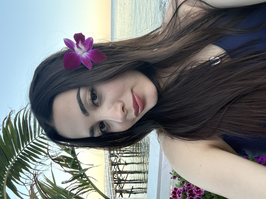 The image depicts a young adult female, positioned in the foreground, taking a selfie or being photographed. She has long, dark brown hair, parted centrally, with a vibrant fuchsia-purple orchid flower pinned above her right ear. Her facial features include dark eyes, defined eyebrows, and lips coated with a reddish-brown gloss. She is wearing a dark blue or navy sleeveless top and a silver-colored pendant necklace.
The scene is set outdoors at what appears to be late afternoon or early evening, indicated by the soft, warm light and subtle orange hue on the horizon. To the left and below the subject, a white railing is visible, with a cluster of purple and magenta flowers in the bottom left corner. Behind the woman, stretching across the midground, is the Pacific Ocean, its surface showing gentle ripples. Prominently in the background, a white wooden pier with numerous support pilings extends out over the water. This structure is identifiable as the Malibu Pier in Malibu, California, United States of America. Above the pier, a few palm fronds are visible, and the sky transitions from a light blue overhead to a pale yellow-orange at the horizon line.