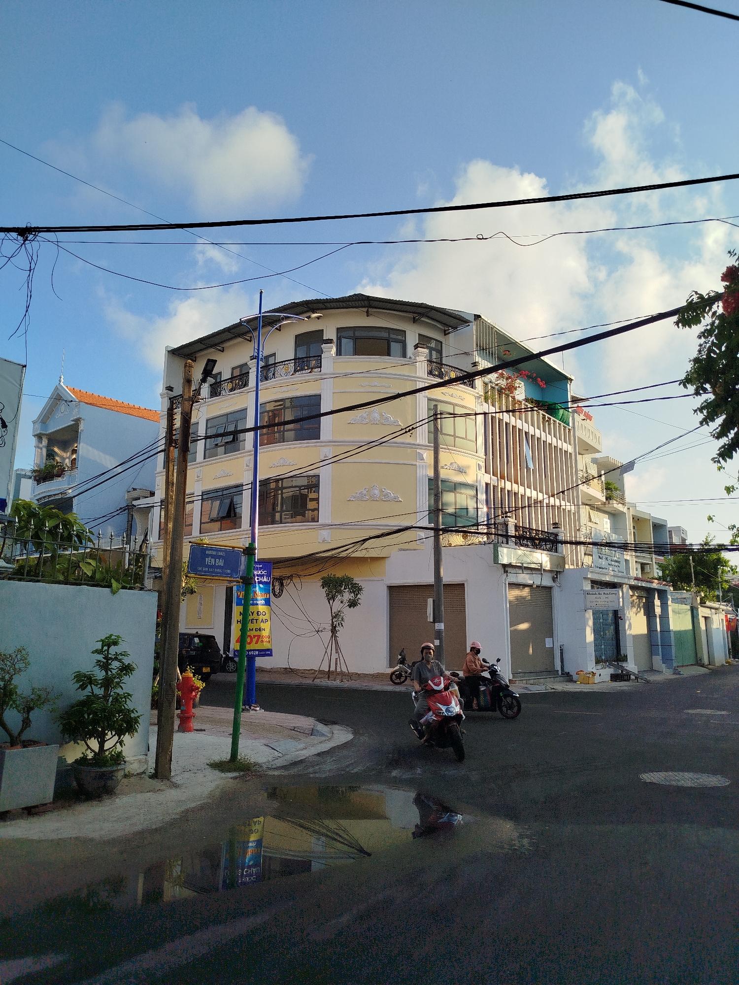 The image depicts a street scene in Ho Chi Minh City, Vietnam. The sky is blue with scattered white clouds. Overhead, there are several electrical wires crisscrossing the sky. The corner of a building with a distinct yellow facade and ornate trim is visible, with a green-and-blue street sign indicating "YÊN BÁI." A red fire hydrant is situated near the sign. Two people on motorbikes are driving on the asphalt road, which has a puddle reflecting the surroundings. The road also shows wear and tear. A variety of buildings are seen along the street, with greenery visible on the left side.
