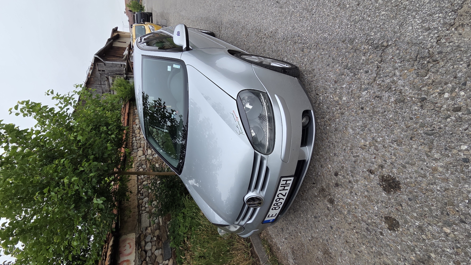 A silver Volkswagen Golf is parked on a grey asphalt road. The car is facing towards the viewer at a slight angle. The front license plate reads "E 8892 HH". There is a small sticker on the hood of the car and another on the driver's side mirror with the letters "skg". To the left of the car is a stone wall topped with red tiles, and behind the wall is a large green tree with dense foliage. Further back, there are old wooden buildings. In the background, other vehicles, including a yellow car and a black car, are parked on the road. The sky is overcast and grey. There are oil stains on the road in front of the car.