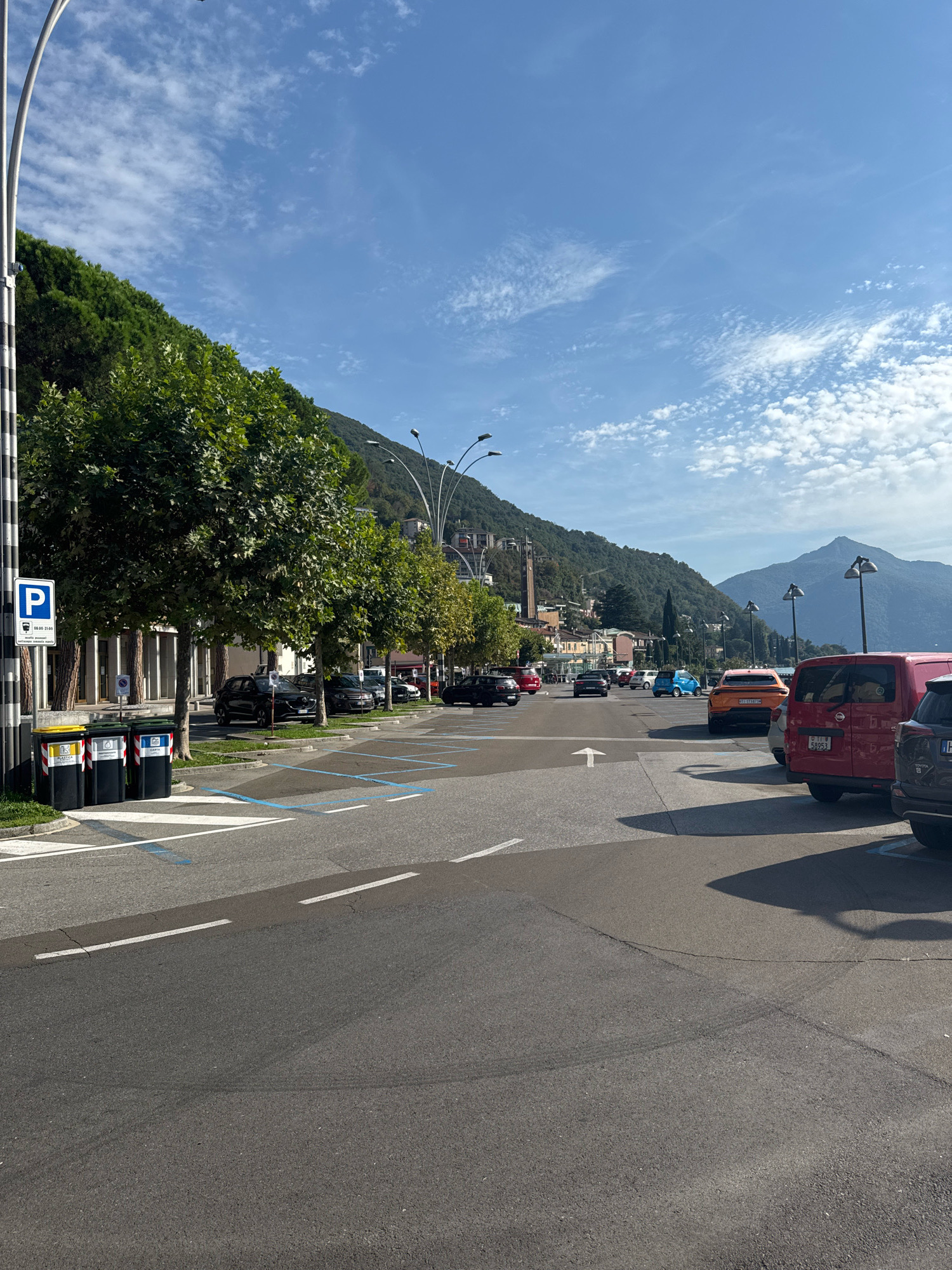 The image depicts a street scene in Campione d'Italia, Italy, characterized by a road with parking spaces marked with blue lines. A tall, modern lamppost with multiple lights stands on the left side of the frame, near a blue and white parking sign. Adjacent to the lamppost are three large trash bins labeled for different types of waste. Several trees with lush green foliage line the sidewalk behind the bins, casting shadows on the pavement. In the background, a hilly landscape covered with trees and buildings rises under a bright blue sky with scattered white clouds. Various cars are parked and in motion on the road, including a red van, an orange car, and a blue car. The overall atmosphere is that of a bright, sunny day in an Italian town.