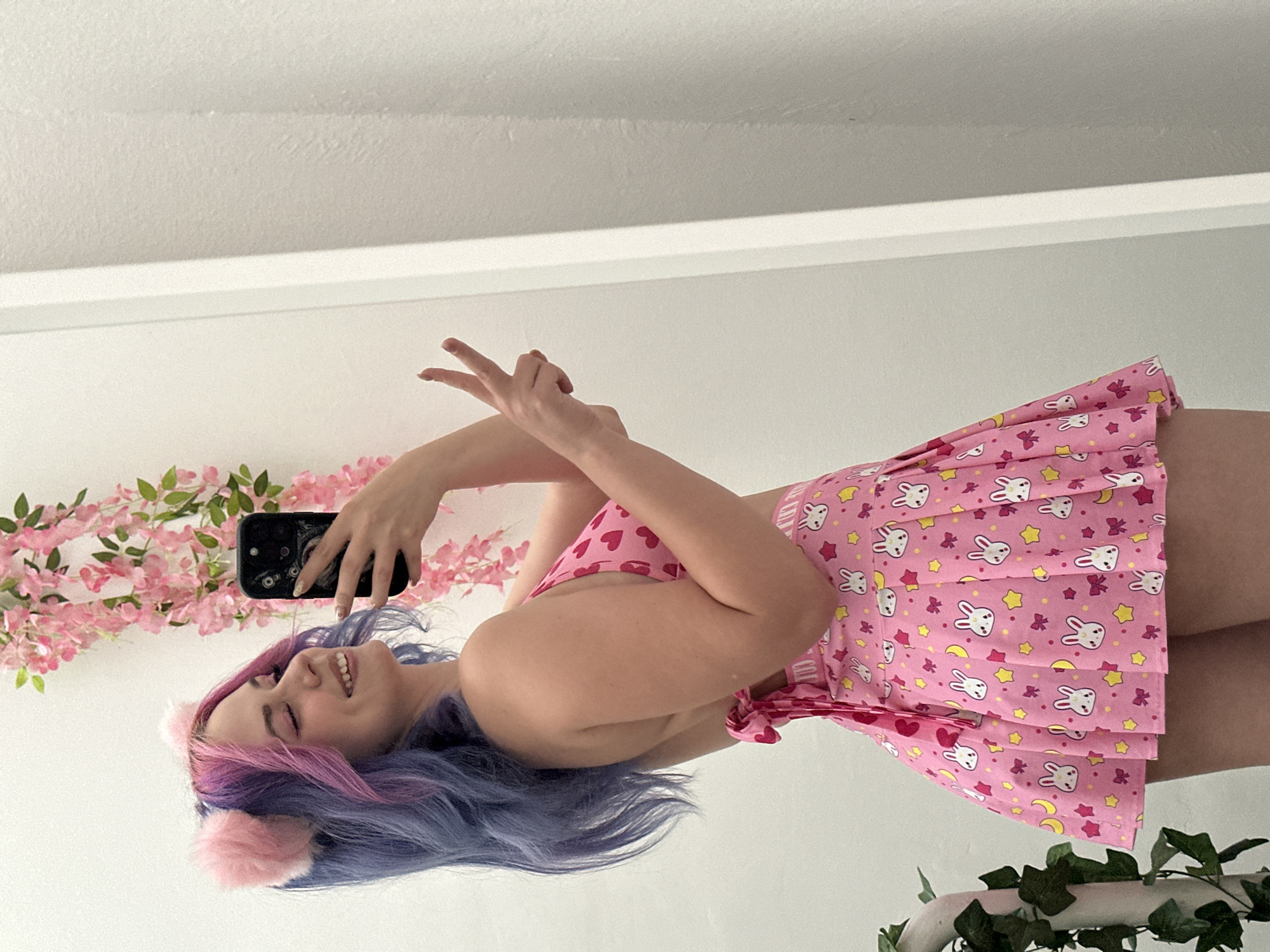 A woman with long, dyed blue and purple hair, adorned with pink cat ears, is smiling and holding a smartphone in her right hand. She's wearing a pink outfit patterned with bunnies, hearts, stars, and moons. A string of pink flowers is visible in the background on the left, while green foliage is visible on the right, likely attached to a white object. The location is likely indoors due to the plain white walls. There is no indication this is in City of Zagreb, Croatia.