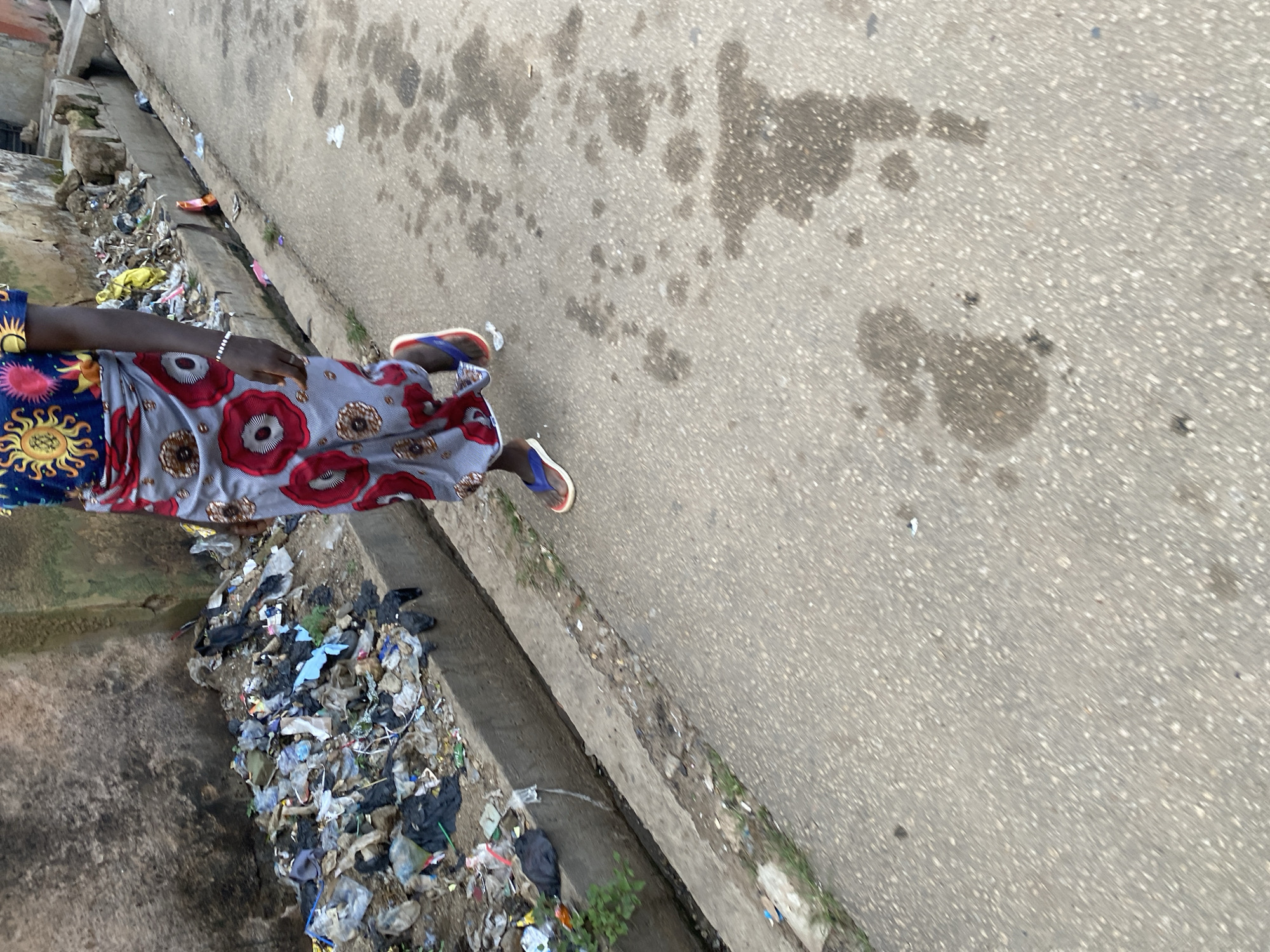 In Jos, Nigeria, a person walks down a street. The person is wearing a colorful patterned skirt and a blue shirt with a yellow and red sun design. They are also wearing blue and red flip-flops. The street is paved with asphalt, and there is a concrete drainage ditch running alongside it. The ditch is filled with trash, including plastic bags, bottles, and other debris. The wall next to the ditch is made of rough, textured concrete. The overall scene depicts a typical urban environment in Jos, with a focus on the contrast between the human activity and the prevalent urban decay.