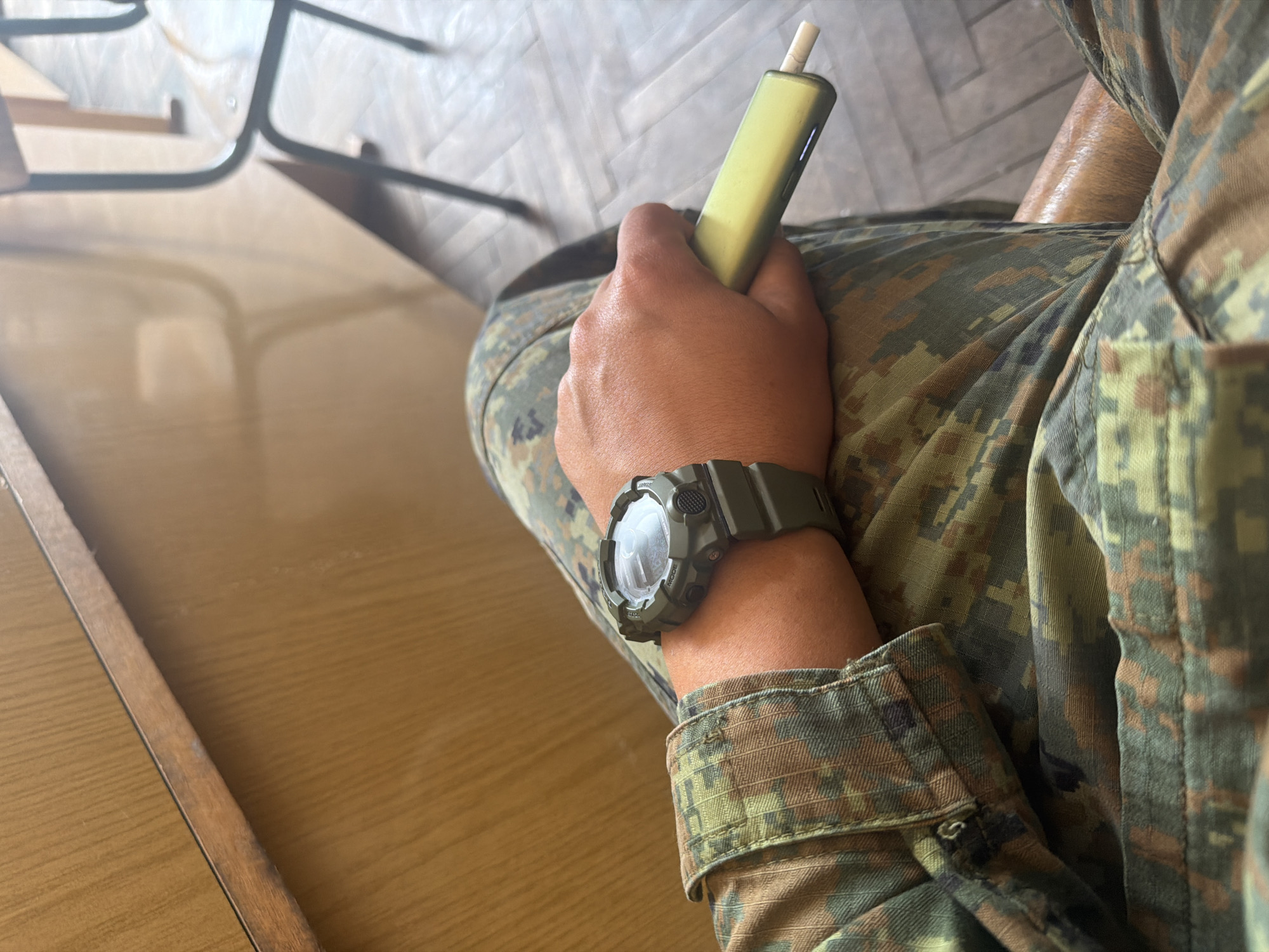 A person in camouflage clothing sits at a light brown table. They are holding a yellow electronic cigarette device. The person is wearing a green digital watch on their left wrist. The background includes a chair and wood floor. No information suggests this is located in Dolna Mitropolia, Bulgaria.