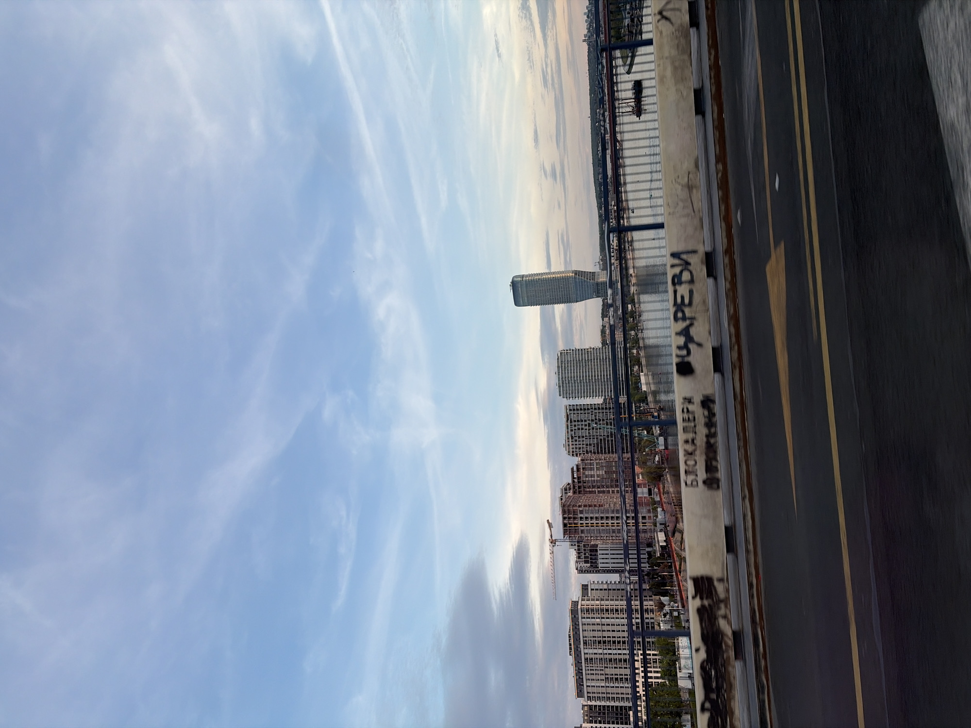 The image shows a view from a bridge, likely in Belgrade, Serbia, looking toward the Belgrade Waterfront development. The buildings are modern, high-rise apartments and office towers. A construction crane is also visible. The foreground includes the bridge's concrete barrier, marked with graffiti, and a section of the road with yellow lane markings. The sky is partly cloudy with a blue hue. The image appears to be taken during the day.