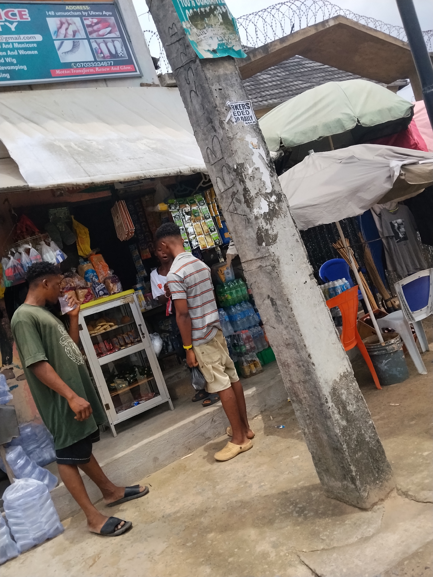 Two young men are interacting near a small outdoor shop in Umuode, Nigeria. One man in a green shirt drinks from a cup, while the other, wearing a striped shirt and khaki shorts, holds a black plastic bag. They stand in front of a shop displaying goods in a glass case and shelves. Behind them, an individual is partially visible inside the shop. An advertisement for "ICE BEAUTY" at "148 umuocham by Ukwu Apu" with the phone number 07033334677 is affixed to an awning. A concrete utility pole covered in posters is prominent in the foreground. Additional merchandise, including bottled drinks, is displayed near the pole and surrounding area. Barbed wire is visible above the shop's roof. Several chairs and a bucket are located adjacent to the storefront.