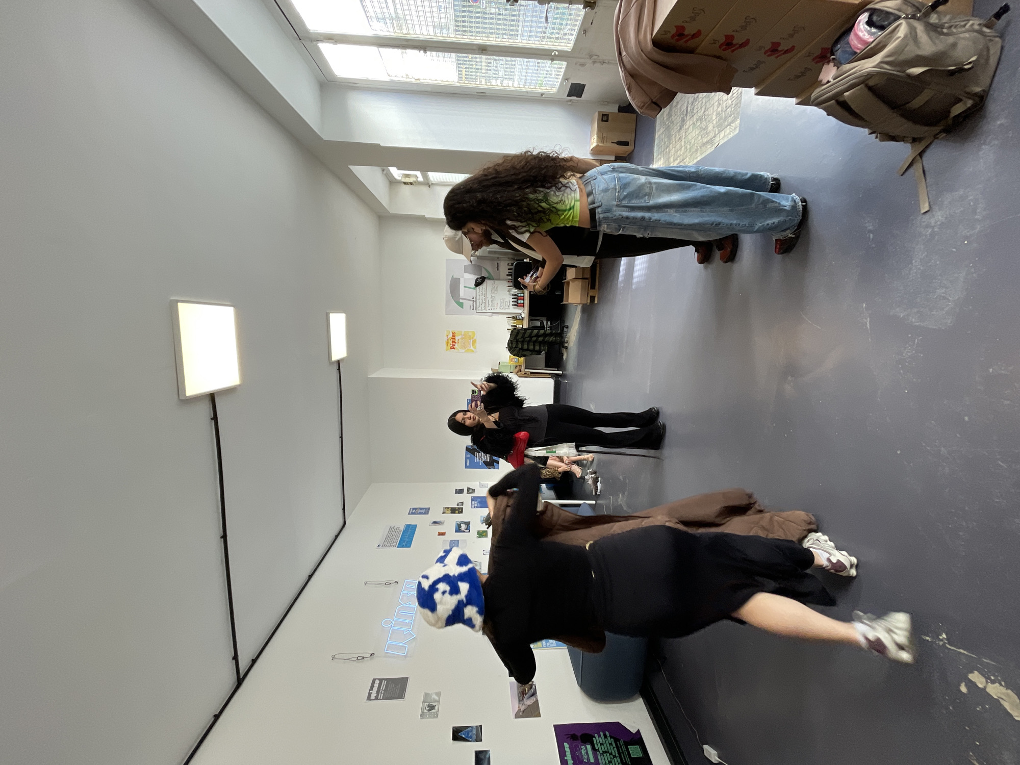 The image depicts an interior space in Paris, France. The scene includes three women. One woman, with long curly hair, wears a light green top and blue jeans, and is bending over towards a table. Another woman is wearing a black outfit and is taking a selfie with her phone. A third woman wearing a black dress, is standing on the right side of the frame and looking to the left. The room has white walls, a gray floor, and large windows at the end of the room. The walls are decorated with posters and signage. There are also light fixtures on the wall. Boxes and bags are stacked in the background.