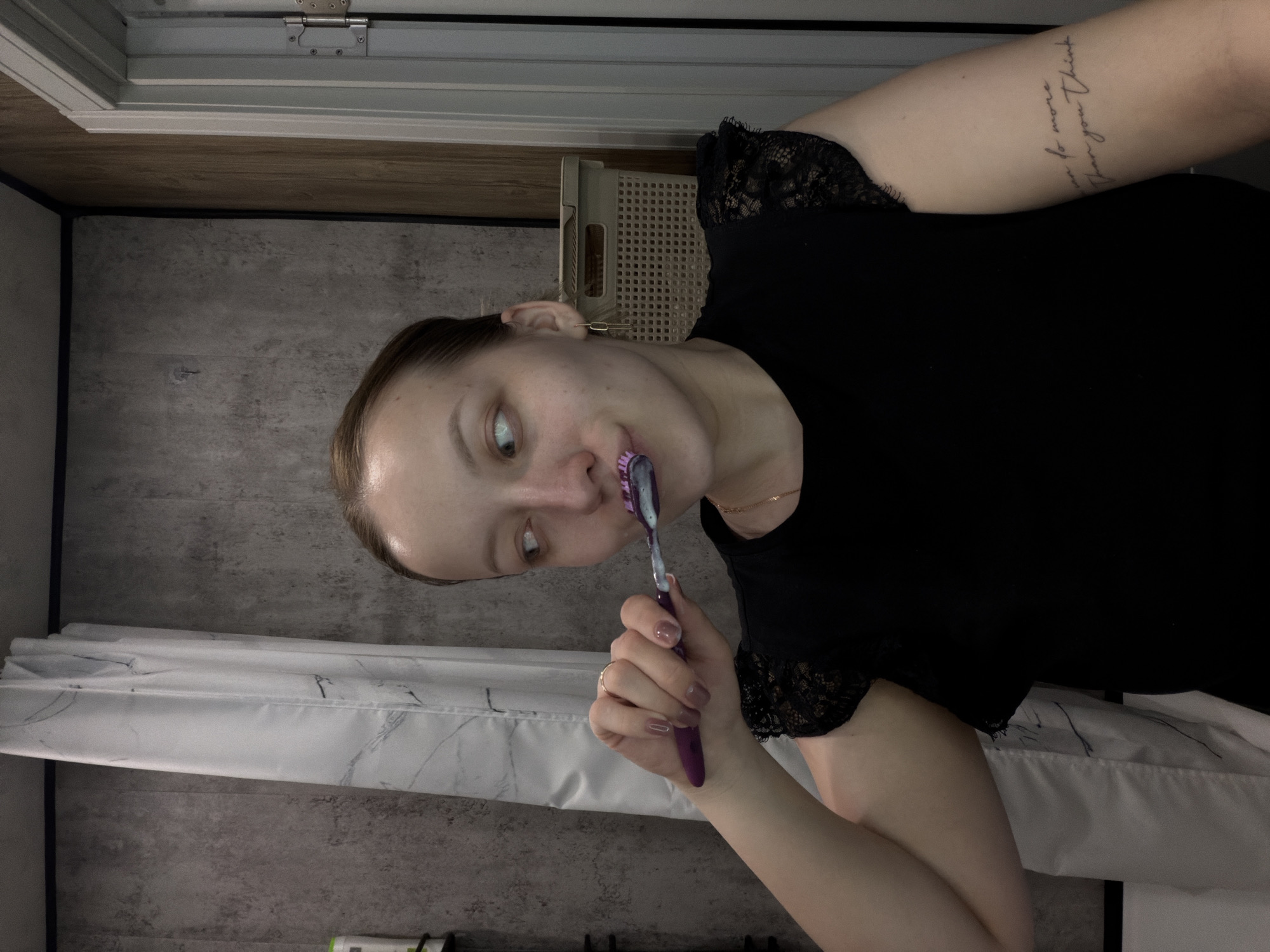 A woman is brushing her teeth in a bathroom. She is wearing a black shirt with lace sleeves. Her hair is pulled back and she has a tattoo on her arm. The bathroom has a grey wall, a white shower curtain with grey marbling, and a white cabinet. There is no information available to determine if the location is Reshetnikovo, Russia.