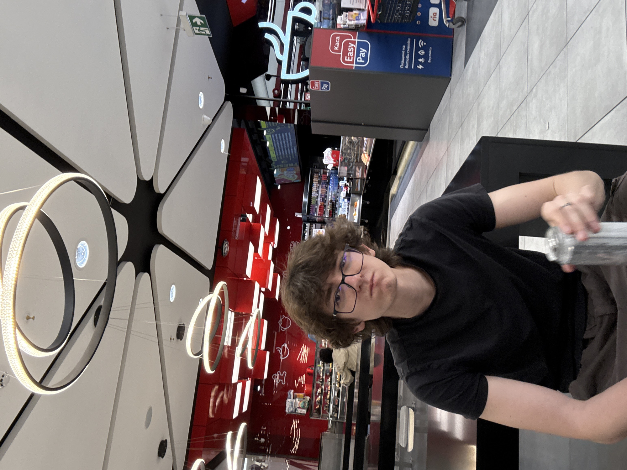 A pale-skinned person with brown hair and eyeglasses is sitting at a table with an aluminum water bottle in their right hand. The person is wearing a black t-shirt. The background includes a modern ceiling design with circular light fixtures, a red wall with white decorative elements, and a “Kasa Easy Pay” sign. This photo was taken in the city of Sofia, Bulgaria.