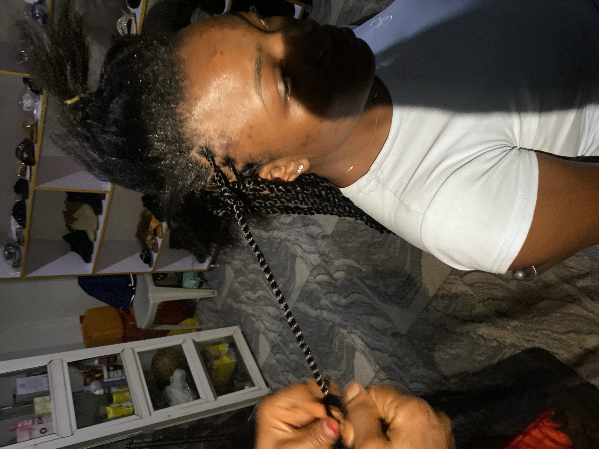 The image depicts an indoor scene, likely a hair salon or a home setting, with elements suggesting a casual environment. In the foreground, a person is having their hair braided. The camera focuses on the upper body and head of the person receiving the service, showing a close-up of the braiding process. One hand, with red nail polish, is visible, skillfully manipulating strands of hair. The background reveals shelves filled with various items, including bottles, boxes, and what appear to be shoes, suggesting a retail or storage area. A large orange container is also present, adding to the cluttered but functional aesthetic of the space. The overall lighting is somewhat dim, with shadows contributing to the atmosphere. The image does not provide specific visual cues to definitively place it within the city of Jos, Nigeria.