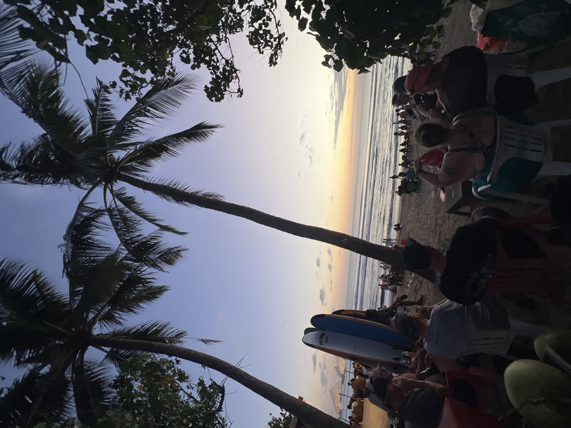 At dusk on Kuta Beach in Kuta, Indonesia, a group of people are seated on chairs on the sand, facing the ocean. Two tall palm trees frame the scene. In the foreground, three surfboards stand upright. The ocean has gentle waves, and several people are visible in the water and walking along the shore. The sky is a gradient of blue and orange, indicating the setting sun. Beach umbrellas and more seating can be seen further down the beach, suggesting a relaxed atmosphere.