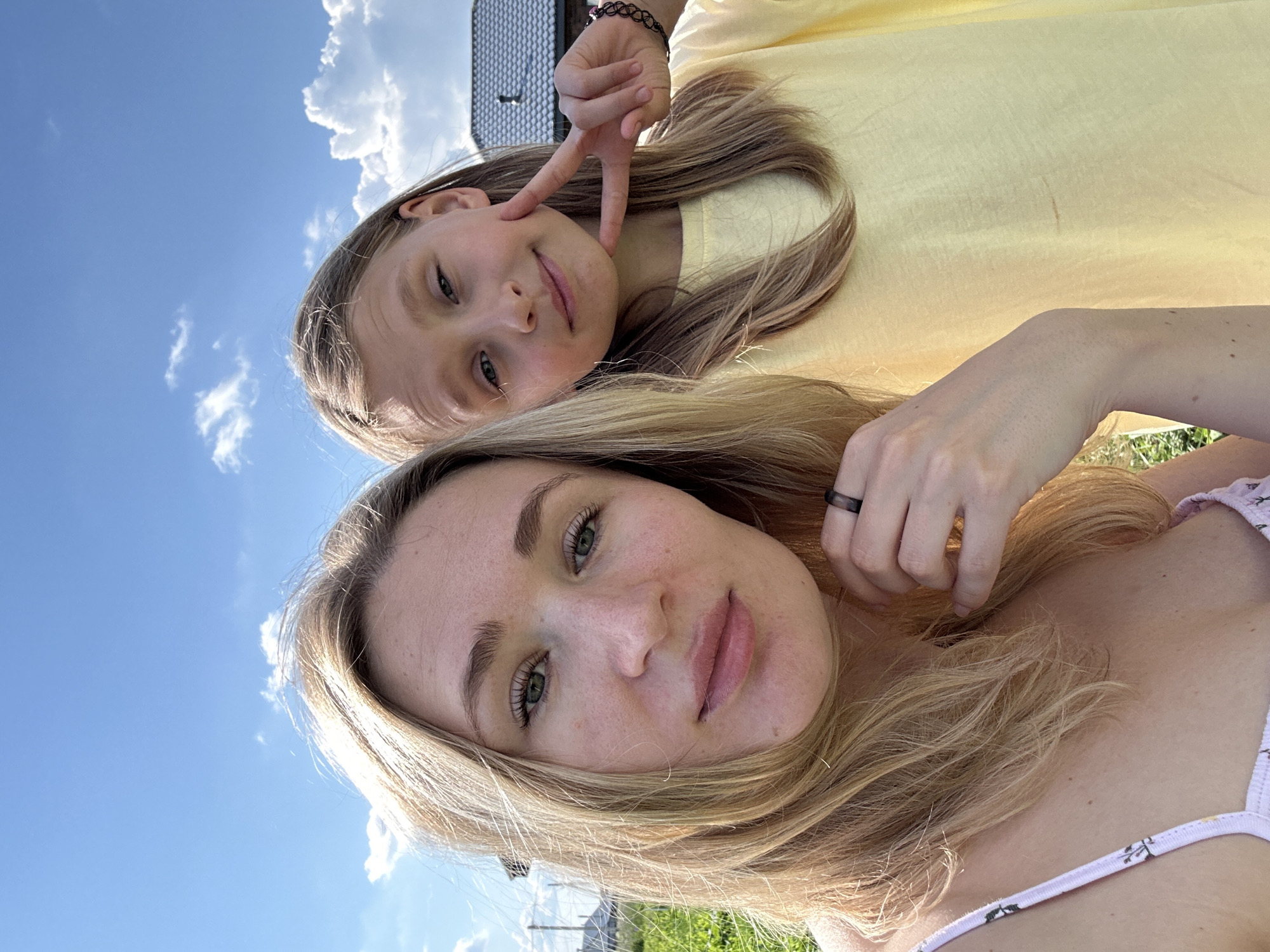 A selfie taken outdoors on a sunny day shows two individuals, likely an adult and a child, in the city of Mokino, Russia. The background features a clear blue sky with scattered white clouds, green grass, and the rooflines of houses, indicating a residential area.
On the left, closer to the camera, is an adult with blonde hair, green eyes, and fair skin. She is wearing a light-colored top with a floral pattern. Her hair is styled loosely, framing her face. She is looking directly at the camera.
To the right of the adult is a younger person, also with blonde hair and fair skin. This individual is wearing a yellow t-shirt and is making a peace sign with their fingers near their face. They are looking towards the camera with a slight smile.
The lighting suggests it is daytime, with the sun casting light on the subjects. The overall scene conveys a casual, happy moment captured outdoors.