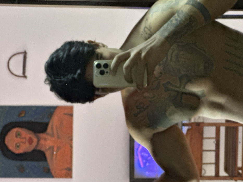 The image captures a mirror selfie of a shirtless individual in an indoor setting, possibly in Guatacaral, Venezuela. The subject, a person with dark, wavy hair, holds a light-colored smartphone (specifically an iPhone with a triple-lens camera array) in their right hand, obscuring the majority of their face. Extensive dark tattoos are visible across the individual's chest and both arms, including a large design on the chest and a solid dark band on the left forearm.
The background, visible in the mirror's reflection, consists of a light-colored wall. Above and to the left of the subject's head, a rectangular painting is mounted, depicting the upper torso of a person with dark hair, green eyes, and orange-toned skin against a dark blue background with abstract, possibly leaf-like, elements. To the right of the painting, a small, curved, brown D-shaped object is affixed to the wall. In the lower-left quadrant of the image, a segment of a wooden furniture item with horizontal slats is visible. Below the painting and adjacent to the subject's left side, a screen (likely a television or monitor) displays a vibrant blue and purple illuminated graphic.