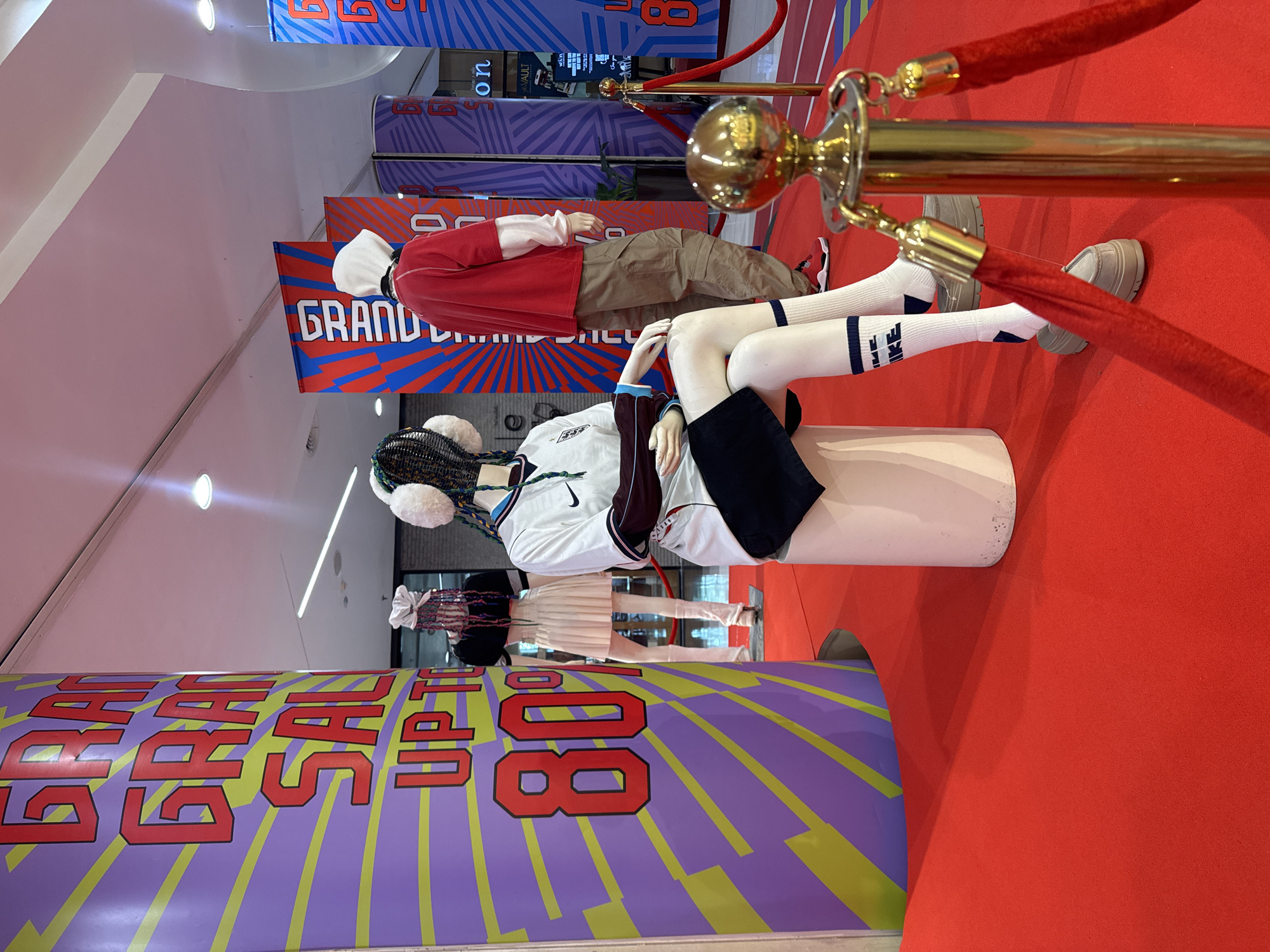 This image depicts a retail store display featuring mannequins in athletic wear, set against a vibrant backdrop of sale advertisements. A large cylindrical pillar on the left is adorned with a "GRAND SALE UP TO 80%" graphic, characterized by bold red lettering on a purple and yellow sunburst pattern. To the right, two mannequins are showcased. The first mannequin, positioned centrally, is seated on a white cylindrical pedestal, wearing a white Nike athletic jersey with dark trim, a black skirt, white socks with navy blue accents and branding, and white sneakers. It has a distinctive hairstyle with colorful braids and white fuzzy ear muffs.
Behind the seated mannequin, a second mannequin stands, dressed in a red long-sleeved shirt layered over a white long-sleeved undershirt, paired with khaki cargo pants and red sneakers. A white hood is pulled up over its head. Further to the right, a third mannequin is partially visible, wearing a white top.
The display is situated on a bright red carpet, creating a sense of urgency and excitement. A gold stanchion with a red velvet rope delineates the display area, adding a touch of premium presentation. In the background, other sale banners with "GRAND SALE" text are visible. The lighting appears to be from overhead fluorescent fixtures, typical of an indoor retail environment. The scene is clean and organized, aiming to attract shoppers. The image does not provide specific indicators of being located in Chiang Mai City Municipality, Thailand; however, the presence of international retail branding like Nike and the general store setup suggest a commercial setting common in urban areas worldwide.