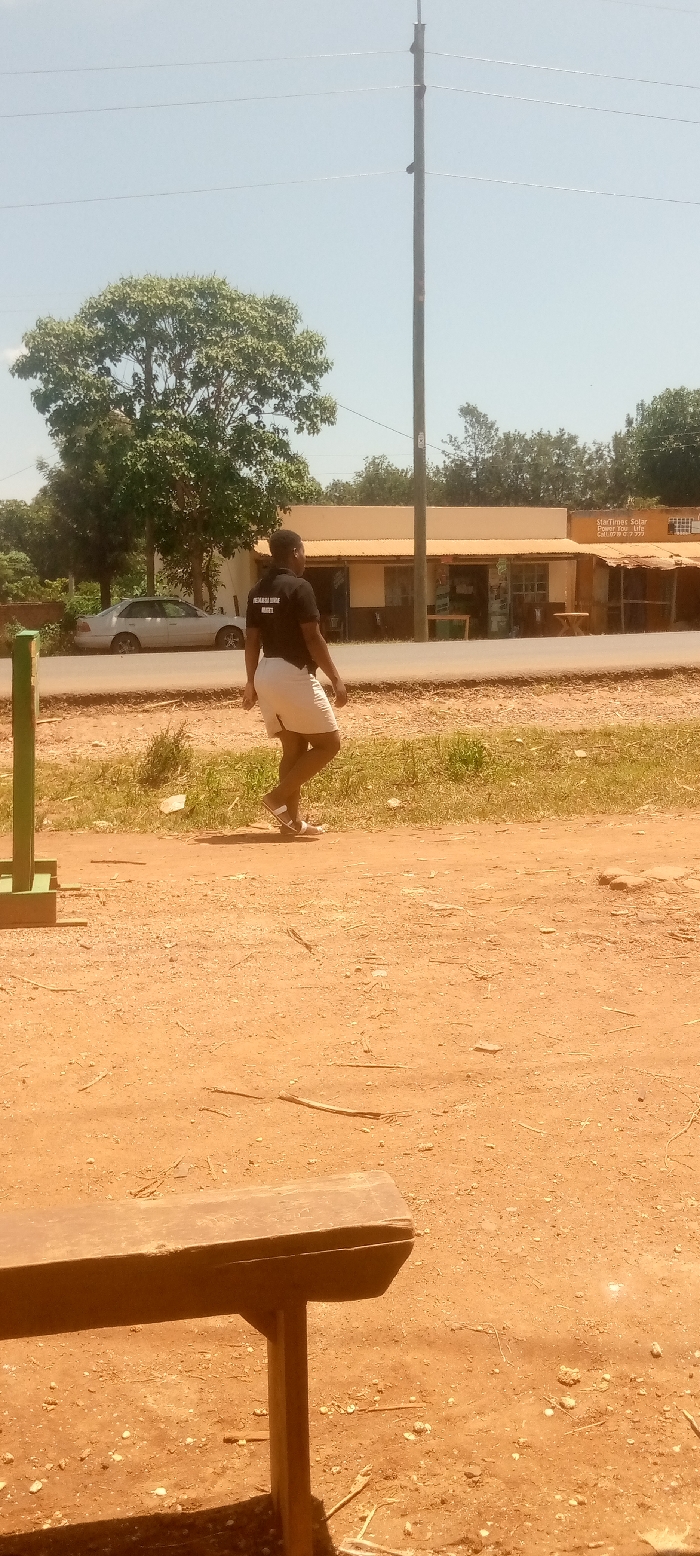 The image shows a man walking on a dirt road in Kivaywa, Kenya. The man is wearing a black t-shirt with a white inscription and white shorts. He is walking away from the camera and towards a small building with a sign that reads "Startimes Solar Products and Services. Call 0700 22 222." There is a wooden bench in the foreground. The man is walking on a dirt road that is surrounded by grass and trees. The sky is clear and blue. The location is latitude 0.56, longitude 34.806 in Kivaywa, Kenya.