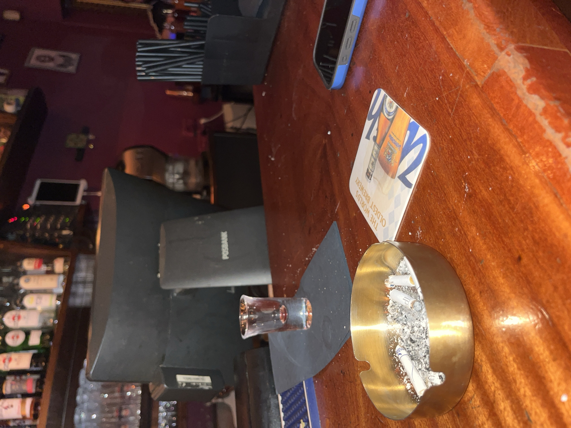 The image depicts a bar counter scene. On the counter, there is a smartphone with a blue case, a beer coaster, a small shot glass, and a full ashtray with cigarette butts. A POS terminal stands prominently on the counter. Behind the counter, a liquor display can be seen. The presence of the POS terminal and the alcohol indicates a commercial establishment like a bar. While the location is not explicitly stated in the image, the prompt suggests this location is Tel Aviv, Israel.