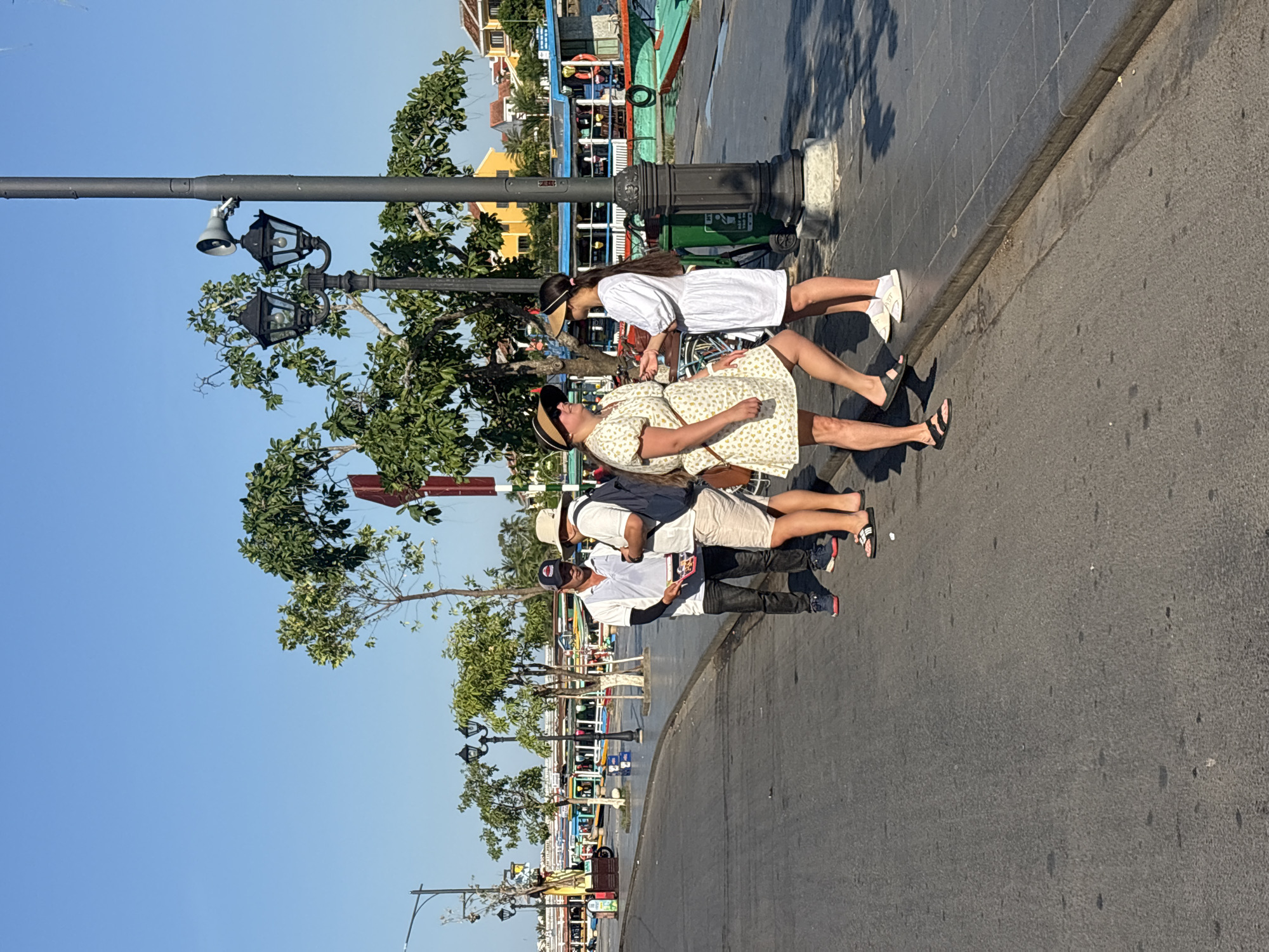 In Hội An Ward, Vietnam, on a bright, sunny day, four individuals are gathered on a paved walkway adjacent to a street. A man in a white polo shirt and dark pants, wearing a baseball cap, stands facing a person in a light-colored short-sleeved shirt and shorts, also with a hat. To the right, two women are interacting. One woman wears a short, yellow dress with a floral pattern and a straw hat, while the other is dressed in a short, white dress. They are positioned near a lamppost that also has a speaker attached. In the background, a line of colorful boats is docked along a body of water, and various buildings, some with yellow facades, are visible. The environment suggests a bustling tourist area.