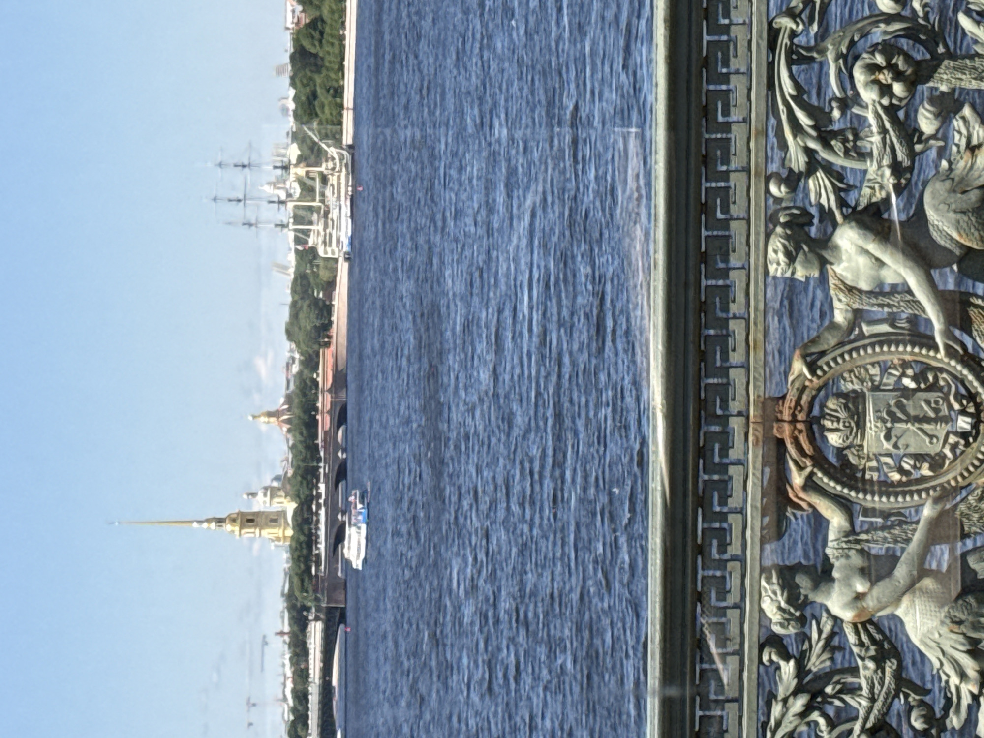 This is an outdoor, daytime, medium shot of a body of water with a cityscape in the background, taken from a bridge railing. The railing in the foreground features decorative metalwork, including two female figures with fishtails and a coat of arms. The water is a deep blue with moderate ripples. In the mid-ground, a white tour boat with people on deck is moving across the water. Further back, a large sailing ship with three masts is docked. The cityscape behind the water features a tall, slender golden spire, several domed buildings, and lush green trees. A bridge with arches is visible crossing the water in the mid-ground. The sky is a clear, light blue. The scene is identifiable as the Neva River in Saint Petersburg, Russia, with the Peter and Paul Fortress in the background.