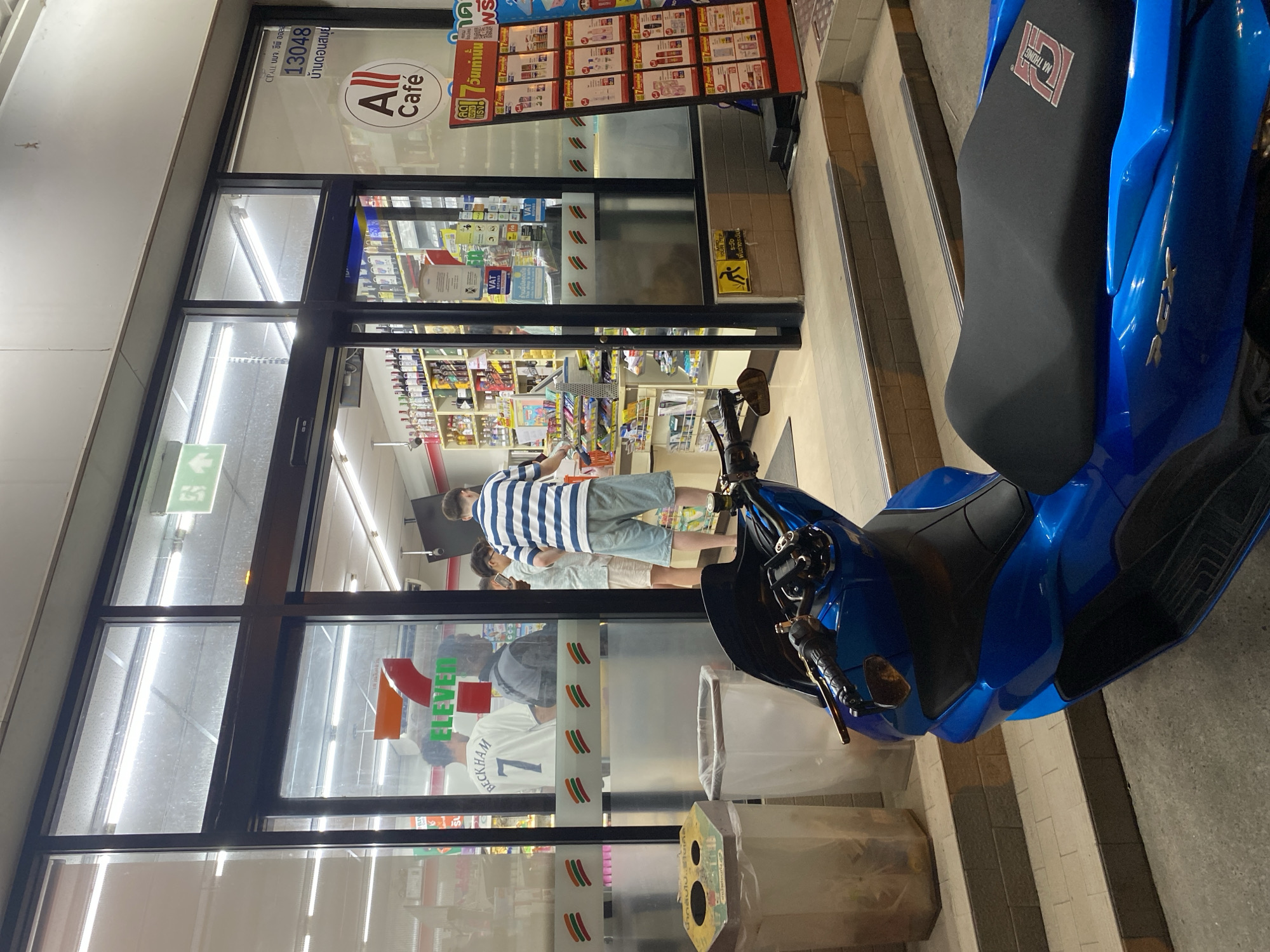 This media file captures an outdoor scene at a 7-Eleven convenience store in Baan Bo Phut, Thailand, likely at night given the artificial lighting. A bright blue scooter is parked in the foreground on the right, its seat prominently displaying a logo that reads "NA THONG."
The storefront is visible, with large glass windows revealing the interior. Inside, shelves stocked with various products are illuminated. Several people are visible through the glass. One man, wearing a striped t-shirt and light-colored shorts, stands near the entrance. Another individual, wearing a white shirt with the number "7" and "BECKHAM" printed on it, is also seen inside. A "7-ELEVEN" logo is clearly visible on the store's exterior.
Text elements within the image include the "7-ELEVEN" branding, "All Café" signage, and Thai text which includes "CPALL บมจ. ซีพี ออลล์" and "13048 บ้านดอนสมุย" (likely identifying the company and the specific store location). There are also yellow warning signs indicating a change in floor level ("พื้นต่างระดับ" and "YOUR DEP"). The overall atmosphere appears to be that of a typical convenience store setting, with the presence of the scooter suggesting it's a common mode of transportation in the area.