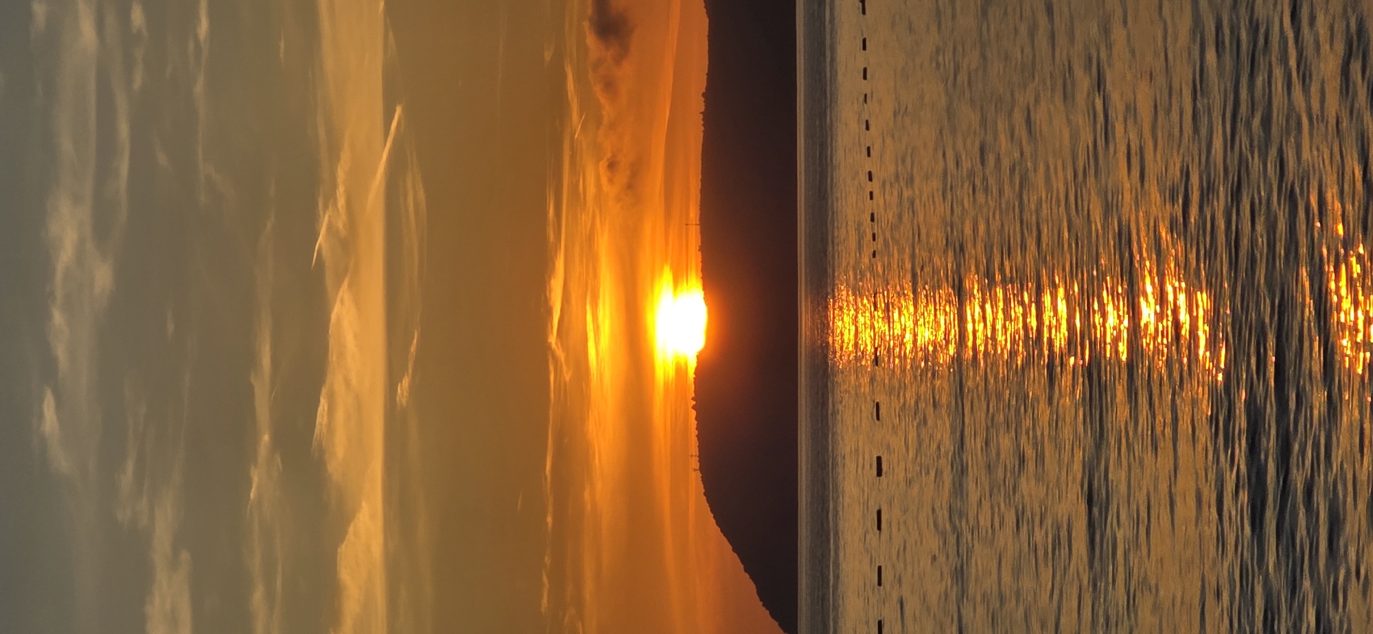The image captures a serene and vibrant sunset (or sunrise) over a body of water, likely the sea, set against the backdrop of Bar, Montenegro.
The upper portion of the scene is filled with the sky, which displays a stunning array of warm colors, transitioning from bright golden and orange tones near the sun to softer, lighter hues with scattered, wispy clouds across the expanse. The brilliant, fiery orb of the sun is a central subject, glowing intensely bright orange and yellow, partially obscured by a dark, silhouetted landmass on the horizon, possibly distant mountains or a coastline.
Below the sun and landmass, the water's surface powerfully reflects the sun's light, creating a dazzling, shimmering path of golden-orange ripples that stretches across the entire width of the frame. The reflection creates vertical streaks of light on the water due to its undulating surface. A notable detail on the water is a series of small, dark, dashed lines, intermittently spaced, suggesting buoys or markers that extend horizontally across the water.
The overall impression is one of peaceful natural beauty during the golden hour, characterized by warm, inviting colors and a tranquil atmosphere. There are no people or explicit man-made structures visible, aside from the potential buoys on the water. No visible text is present in the image.