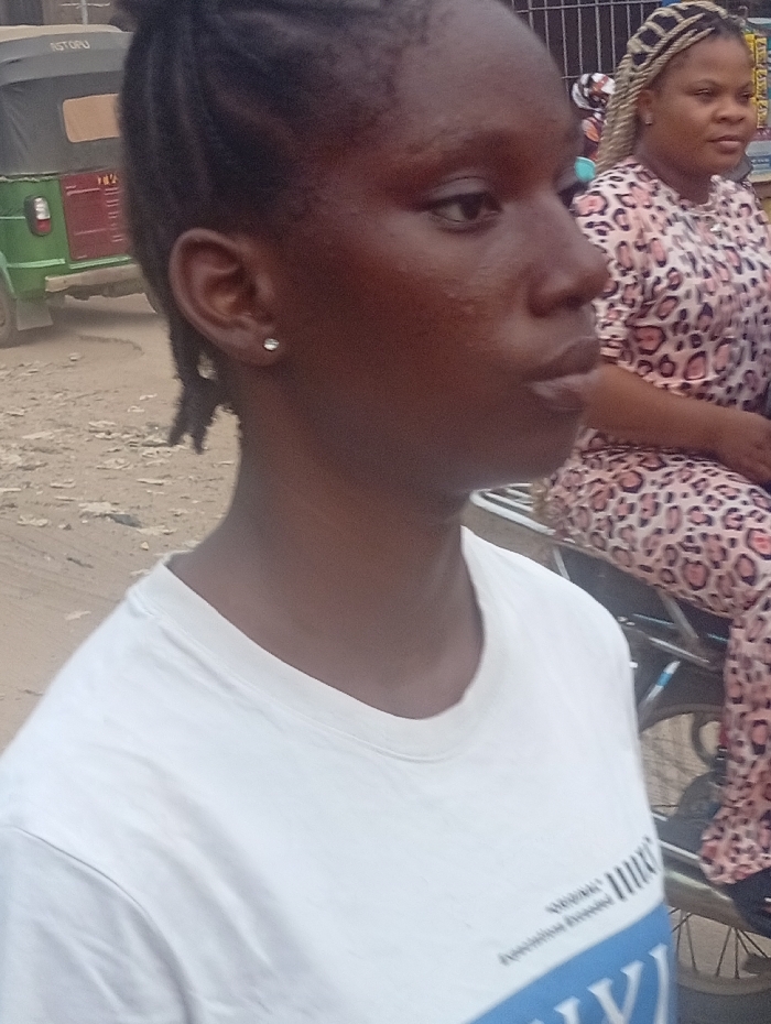 The image captures an outdoor scene, likely a street or public area during the daytime, featuring two women.
In the foreground, a young woman with dark skin is prominently featured in a profile view, facing towards the right side of the frame. Her hair is styled in dark braids pulled back from her face. She wears a small, sparkling stud earring in her left earlobe. Her skin has some visible texture, particularly on her cheek. She is wearing a plain white round-neck t-shirt. Visible text on her t-shirt, though partially obscured, includes what appears to be "American Dream" and smaller text below it that might be "Feminist Press" or similar. Below these, large blue letters, possibly "VY" or "WY", are visible at the bottom of the shirt.
In the mid-ground, to the right and slightly behind the foreground woman, another woman is visible. She has lighter skin and blonde braided hair. She is wearing a patterned outfit, which appears to be a leopard or cheetah print dress or top and pants. She is also facing towards the right but is slightly more angled towards the viewer, and appears to be seated, possibly on a motorbike or scooter, part of which is visible in the lower right.
In the far background on the left, a green and red motorized rickshaw (commonly known as a Keke Napep or auto-rickshaw) is parked on what appears to be an unpaved or dirt road. The word "ASTORU" is visible in white letters on the side of the rickshaw. Behind the background woman, a metal fence or gate is visible. The overall impression is a casual, everyday street scene.