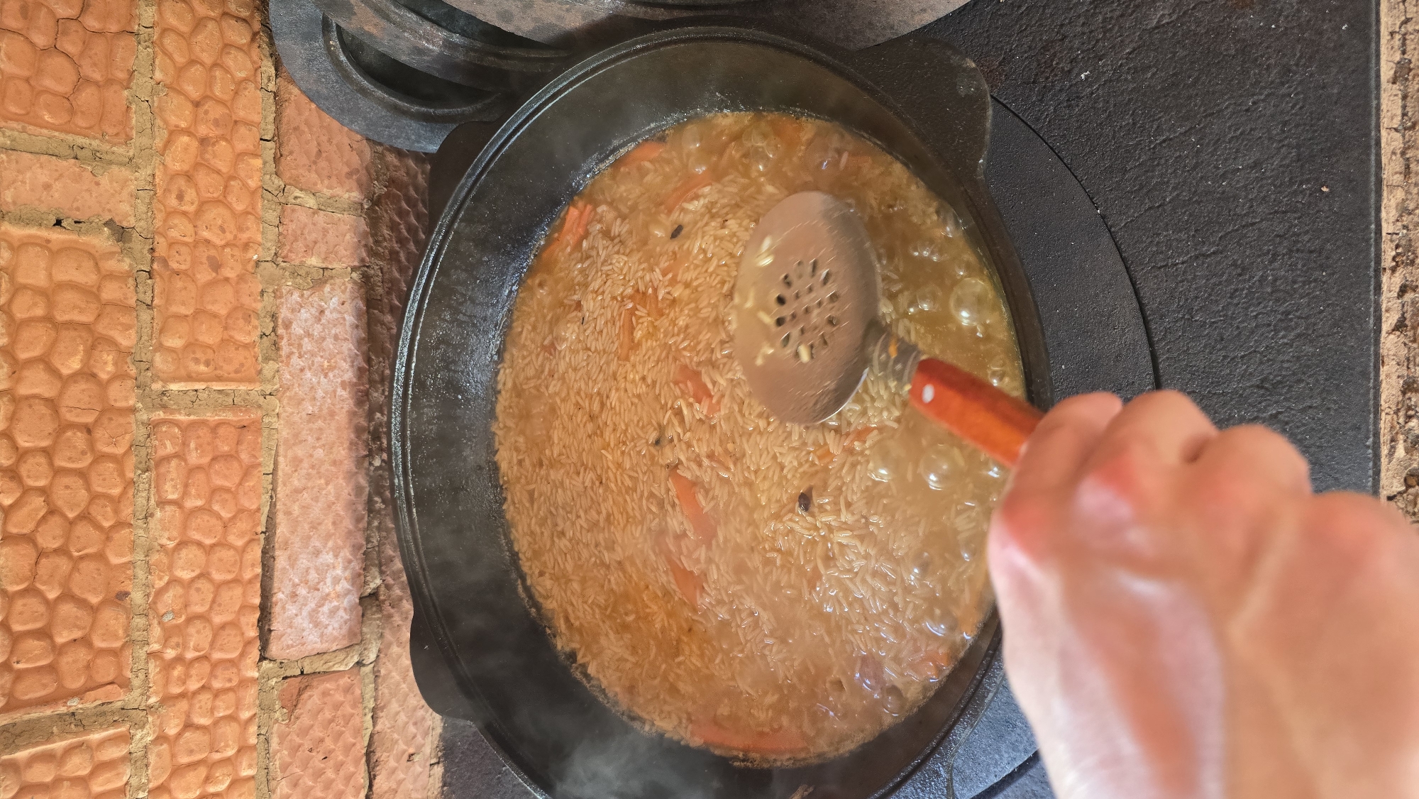 In the city of Забегалово, Russia, a close-up, elevated perspective shows a large, dark cast-iron kazan pot centrally positioned over a stove. The pot contains a light brown rice dish, intermingled with distinct orange carrot pieces and dark spices, simmering in a reddish-orange liquid, actively bubbling with visible steam rising. A fair-skinned human hand is visible in the lower-left, holding a metallic slotted spoon with a red-orange wooden handle, the spoon partially submerged in the contents of the pot. The kazan rests on a dark, textured, circular cast-iron stove surface; additional circular cast-iron rings are stacked on the stove to the right. The background features a rustic wall composed of irregularly shaped, reddish-orange bricks with visible, uneven mortar lines.
