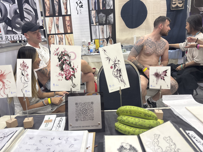 This image depicts an indoor scene at a tattoo convention or studio booth in Ban Rong Ruea Kham, Thailand. In the foreground, a display table showcases various tattoo designs. Prominently displayed are four upright samples: a red floral design, a black dragon amidst pink cherry blossoms, a bamboo motif with a red sun, and a purple butterfly. Below these, several flat sheets present smaller, intricate designs, including butterflies, flowers, and celestial symbols. A stand featuring a QR code linked to "INS:DAVID.GEORGE.TATTOO" is positioned centrally on the table. To the right, three green, elongated objects resembling gourds or cucumbers, inscribed with text, are stacked.
On the left side of the frame, a male wearing a black baseball cap and a white t-shirt is seated next to a female with long hair, dressed in a light-colored top. Both are observing the activity to their right. On the right side, a shirtless male with extensive tattoos covering his chest, arms, and legs is seated in a chair, looking towards another individual positioned further to the right. This individual, with long hair, wearing a light-colored top and dark pants, is facing the tattooed male, appearing to be engaged in a discussion or preparation for a tattoo session. The background features multiple banners, including a black and white snake design on the left and a cream-colored fabric banner with a dark circular design and Japanese characters on the right. Tattoo equipment and supplies are visible on a table behind the shirtless male.