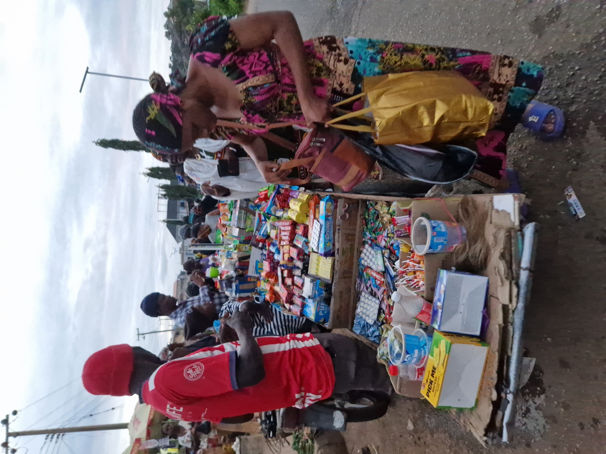This image depicts a street scene in Kabong, Nigeria, possibly a market area. In the foreground, a woman wearing a colorful, patterned dress and headscarf is engaged in activity near a stall. She is holding two bags, one a golden tote bag and another smaller, reddish-brown bag. Her attention appears to be directed downwards.
Adjacent to her is a mobile vendor's stall, laden with various packaged goods, including candies, snacks, and what appear to be toiletries or small household items. The stall is made of wood and appears to be on wheels, suggesting it is a portable setup. Several people are visible in the background, some appearing to be customers and others vendors, engaged in their daily activities. A red t-shirt with "IFE" is visible on a man in the left part of the image, who is standing near a motorcycle. The overall environment suggests a bustling urban setting, with a paved road and overcast sky.