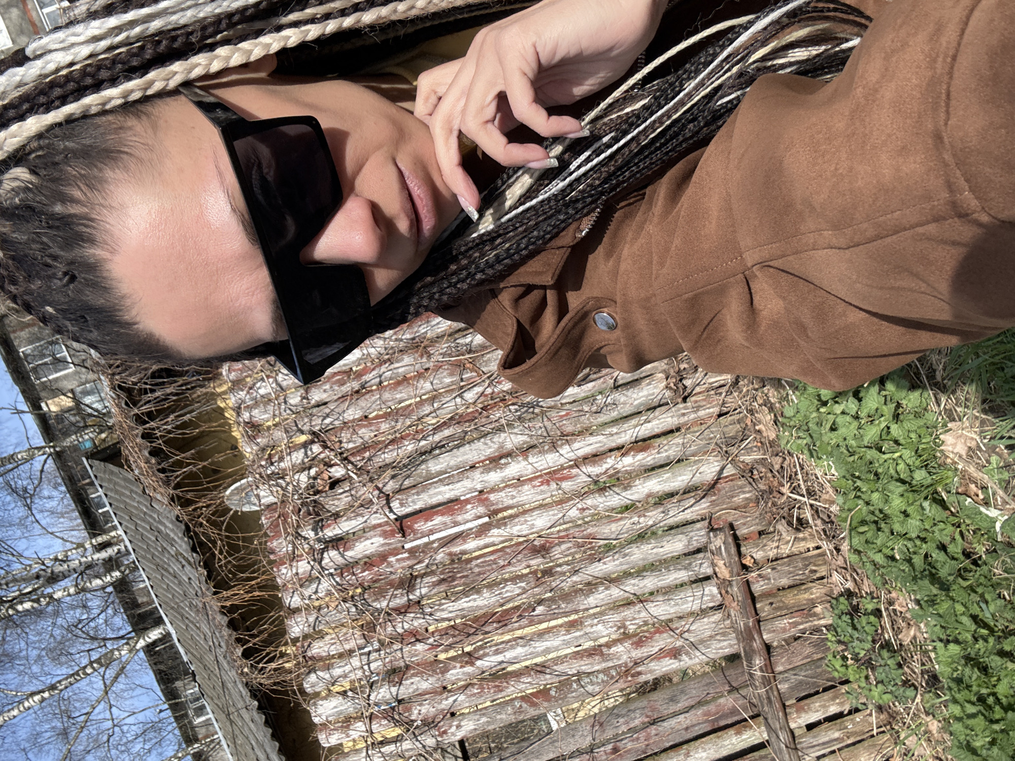 A medium shot depicts a person wearing braids and large black sunglasses, dressed in a brown jacket. They are lying on a weathered wooden surface covered in dry vines. To the side, there is green vegetation and a glimpse of a building with bare trees in the background, possibly in a park or rural setting. The scene potentially suggests Rusanovo, Russia, given the context of the request.