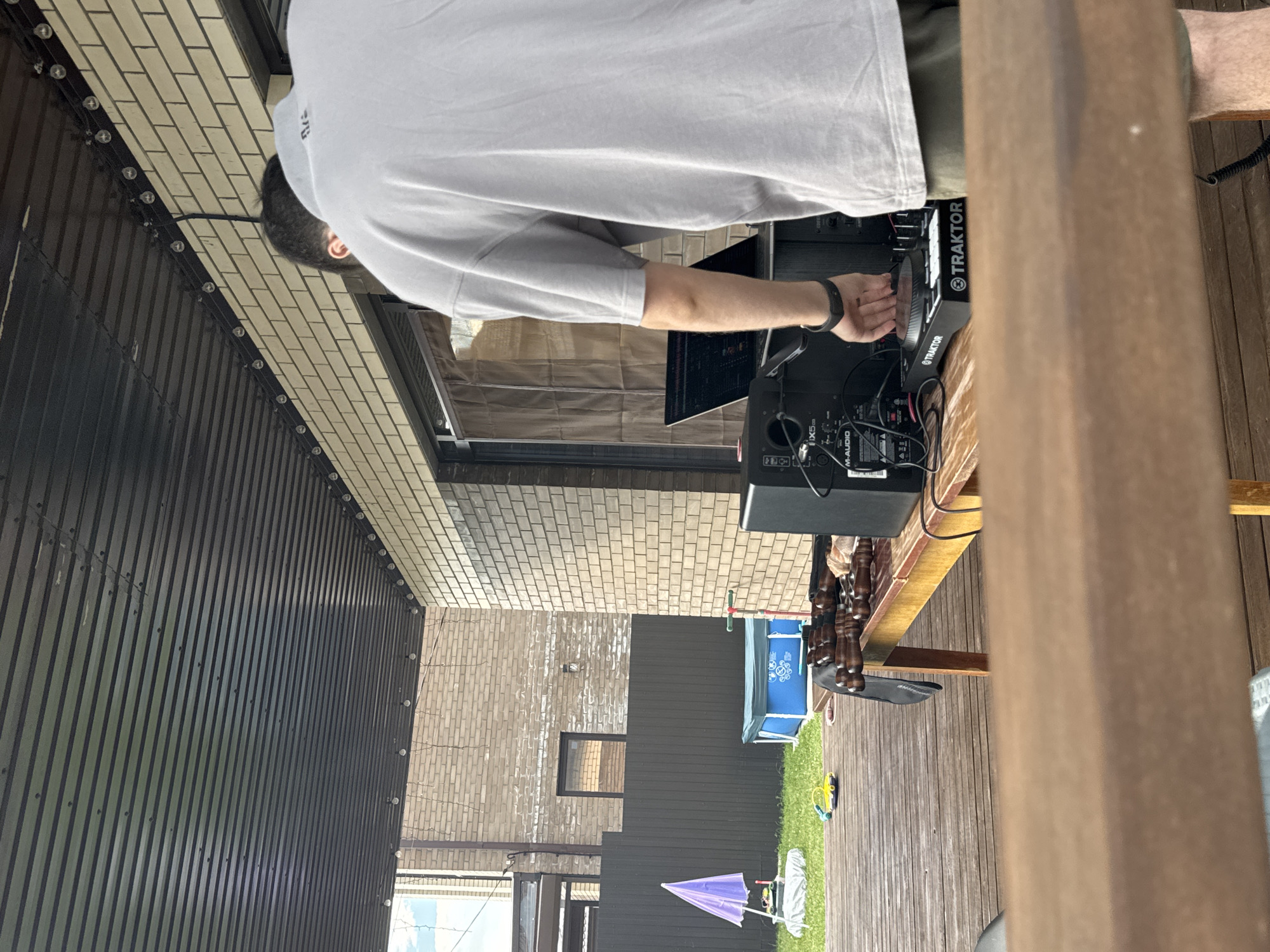 The image depicts a person, likely a male based on attire, engaged with DJ equipment on a wooden deck, possibly outdoors. The person is wearing a grey t-shirt and olive green shorts and has a black wristband on their left wrist. They are facing away from the camera, with their hands over a DJ controller labeled "TRAKTOR". A laptop is positioned behind the controller, and a speaker is to the left of the equipment.
The environment suggests a backyard or patio setting. A wooden table, partially visible, is positioned in front of the DJ setup, with what appears to be skewers or grilling utensils on it. In the background, a dark fence encloses a grassy area with a small inflatable swimming pool and a purple umbrella-like object. A brick building with a dark-colored roof is visible to the right and behind the DJ. The overall lighting suggests daytime. The location is identified as Дёмино, Russia.