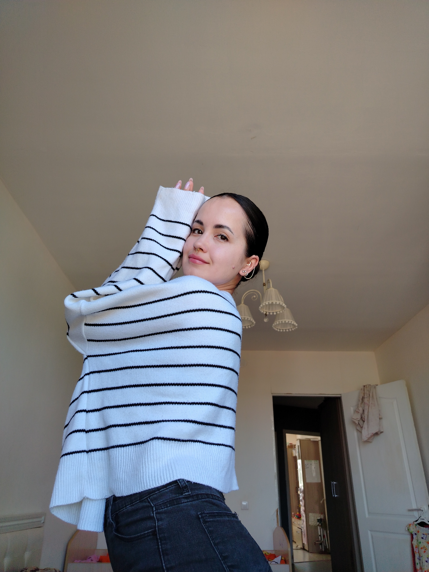 A young woman is posing indoors. She is wearing a white sweater with black horizontal stripes and dark gray jeans. She has dark hair pulled back, multiple earrings in her left ear, and a nose ring. Her arms are raised near her head. The room has light-colored walls and a ceiling light fixture with multiple shaded lamps. An open door reveals another room with visible items and furniture. A partially open white door is visible in the background on the right. There is no evidence to suggest the location is Noviy, Russia.