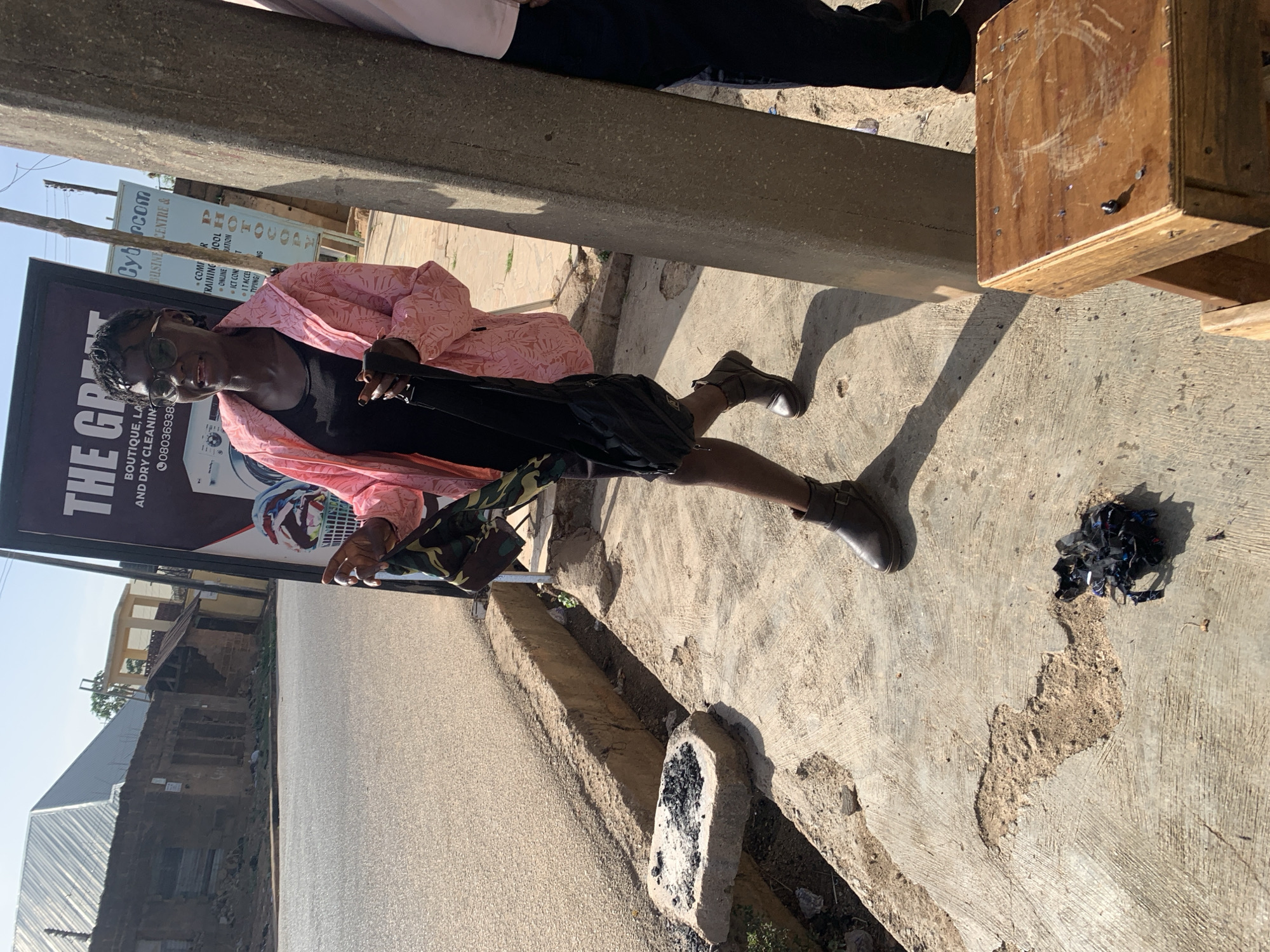 A person wearing a pink shirt, black shirt, and black shorts stands outside in what appears to be Jos, Nigeria. They are wearing boots and glasses and holding a black object. Behind them is a sign for a laundry and business center, as well as a street and buildings. Beside the person is a wooden box and a concrete divider. There is debris on the ground beside them. The environment seems to be an urban area with signs of construction or repair.