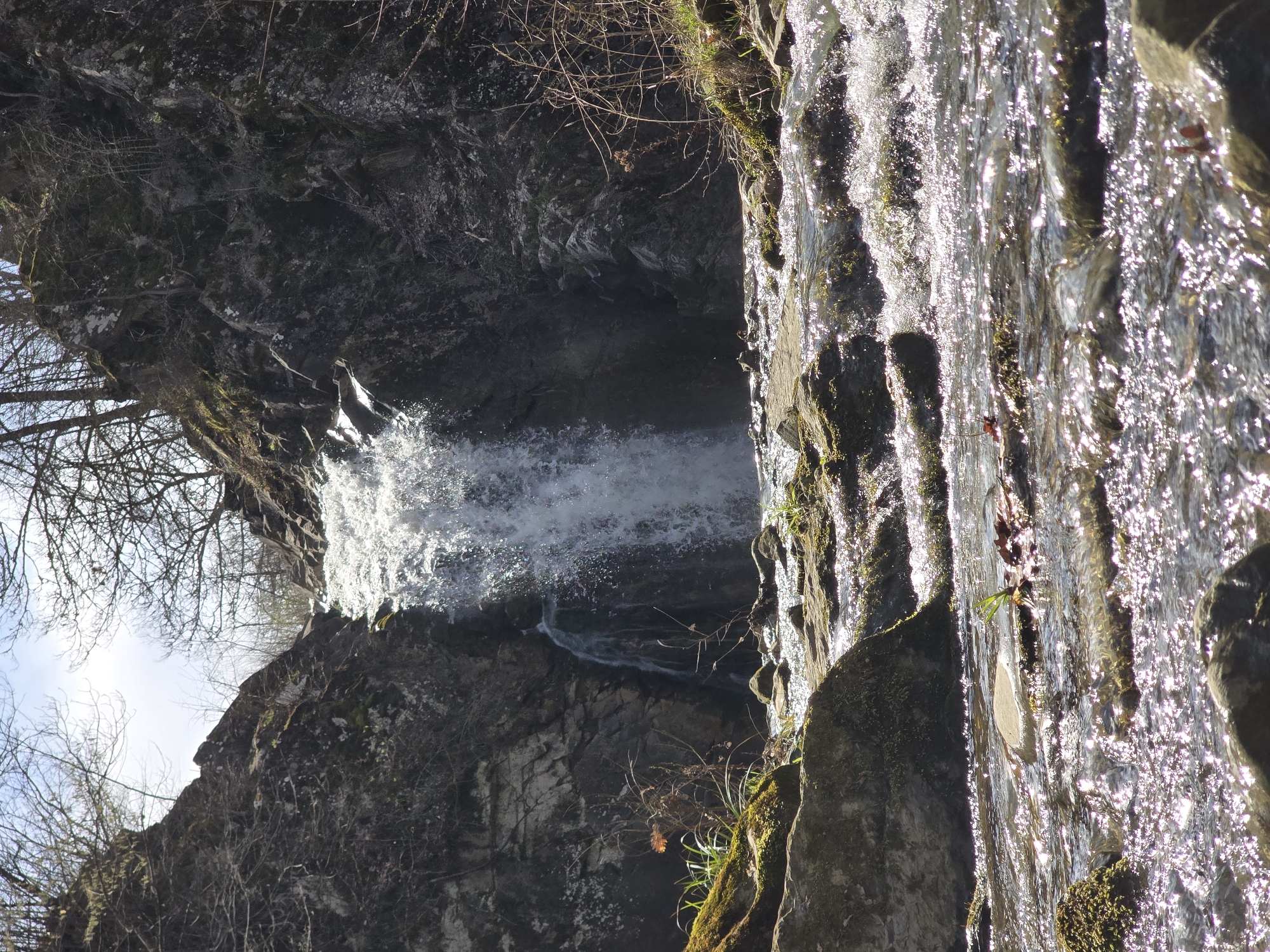 The image depicts a waterfall cascading down a rocky cliff face. The water appears white and frothy, contrasting with the dark, moss-covered rocks. Bare tree branches are visible in the upper left corner of the image. The location is Unknown, Unknown.