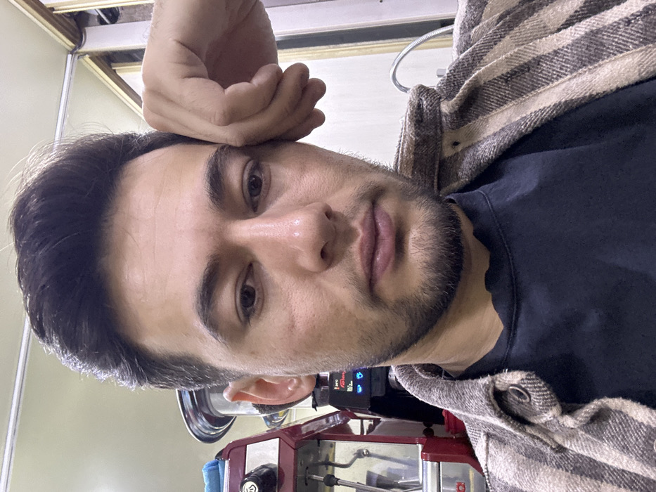 A person rests their head on their hand, looking directly at the camera. They have dark hair and a beard. The person wears a black shirt underneath a beige and grey patterned jacket. An espresso machine is visible in the background to the left of the subject. The background includes a wall with a light fixture.