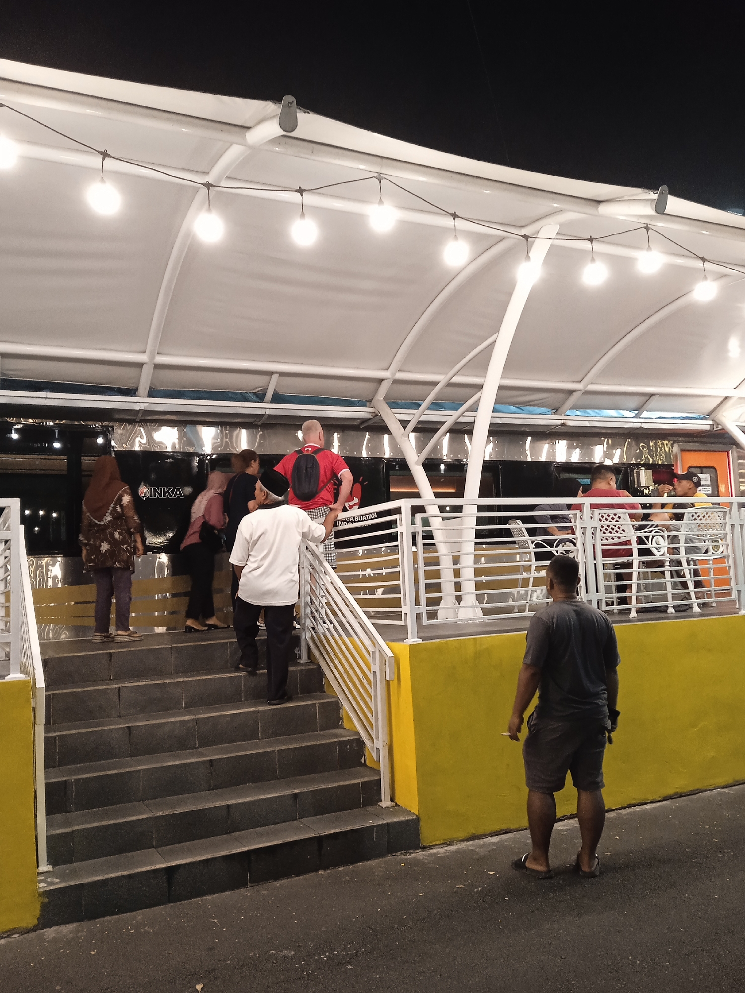 This nighttime image captures a lively outdoor or semi-outdoor dining area, likely a cafe or restaurant, in Madiun, Indonesia. The setting features a white canopy supported by white beams, adorned with string lights that illuminate the space and create a warm ambiance against the dark sky. A prominent yellow wall forms the base of the structure, leading up to a set of grey concrete stairs with white railings.
Several people are present, indicating an active scene. In the foreground, a man in a dark grey t-shirt and shorts, wearing sandals, stands with his back to the camera, holding what appears to be a cigarette. A group of individuals is seen ascending the stairs towards the main seating area. This group includes a woman in a brown patterned top and head covering, a person in a light pink top, a man in a white shirt and black peci (cap), and another man in a red t-shirt with a black backpack.
Behind the white railing, inside what looks like a seating area, at least two men are visible seated at a white table, one in a red shirt and another wearing a yellow cap. The seating area is adjacent to a dark, reflective surface resembling the side of a train car.
A significant detail is the visible text "INKA" on the side of the train-like structure. This refers to PT Industri Kereta Api, Indonesia's state-owned rolling stock manufacturer located in Madiun, strongly indicating the local context. Partially visible text to the right reads "A BUATAN INDO," likely part of "Kereta Api Buatan Indonesia" (Indonesian Made Train), further confirming the train theme. The scene suggests a casual evening gathering or dining experience at a location potentially themed around or adjacent to a train.