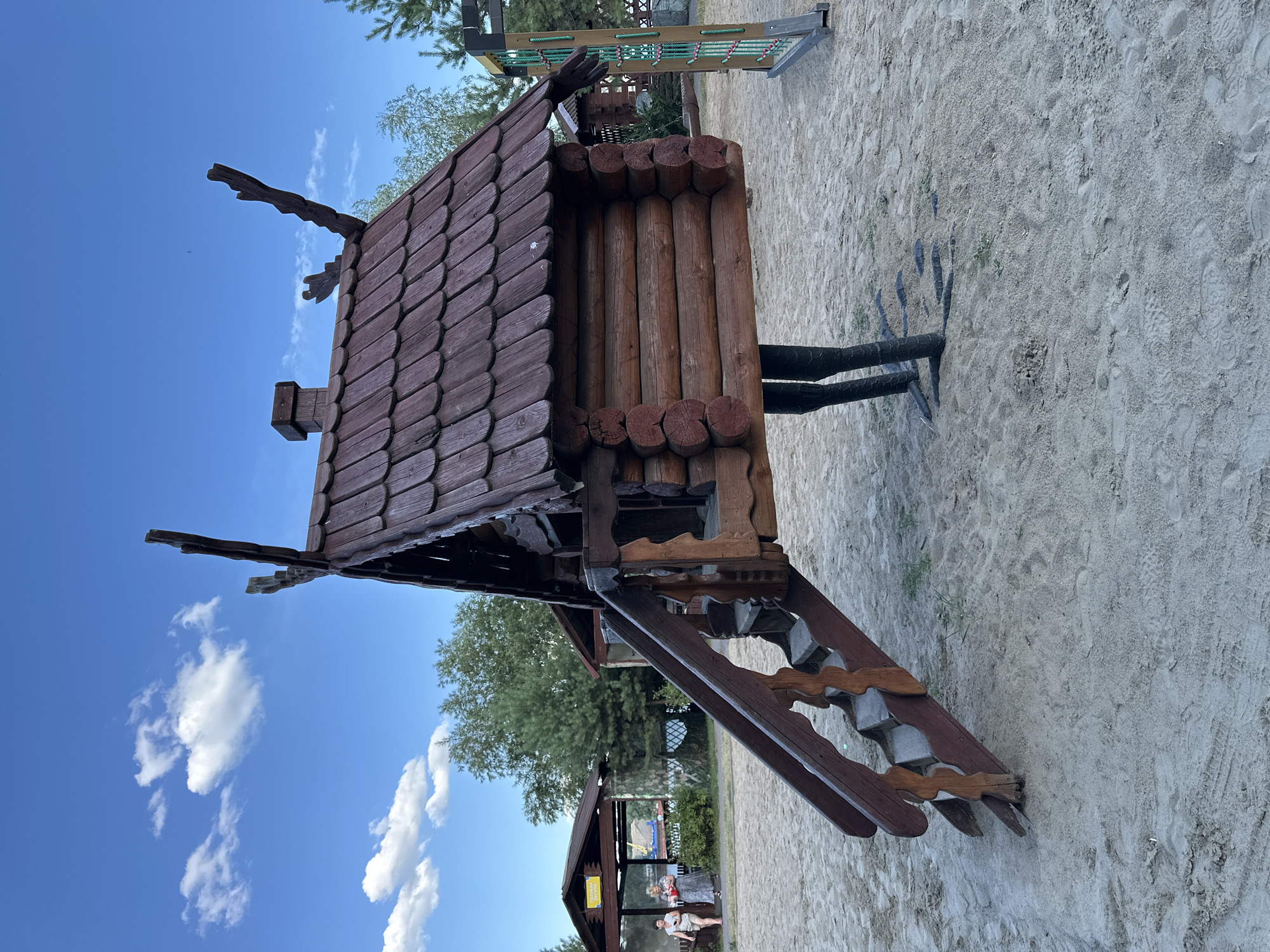 The image depicts a wooden playground structure resembling a log cabin with a pitched, shingled roof, situated on a sandy surface in what appears to be an outdoor recreational area. The cabin is elevated and supported by dark, stylized legs. A wooden staircase with decorative side panels leads up to an opening in the cabin. In the background, there are trees, some covered structures with awnings, and a blue sky with scattered clouds. To the right, a section of another playground apparatus with green rope netting is visible. Two women are present in the background, standing under one of the covered structures. The scene is consistent with a park or amusement area, and while no specific landmarks are visible, the presence of a stylized log cabin suggests a theme park or family entertainment center, possibly inspired by Russian folk architecture. It's important to note that the provided image alone does not contain explicit information to confirm the location as Omsk, Russia, or any specific city.