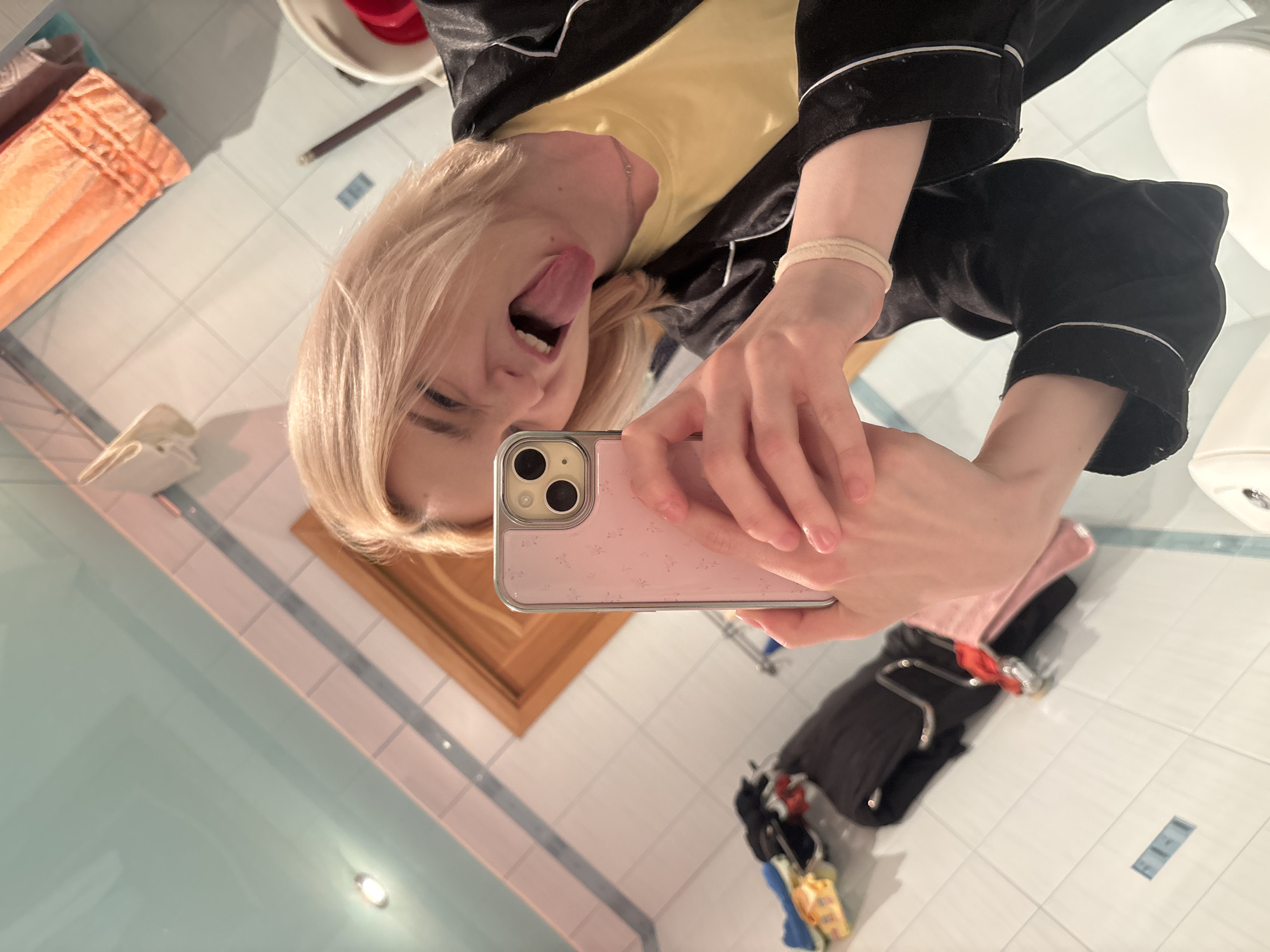 An overhead shot captures a young person in a bathroom, taking a selfie with their phone. They have short, blonde hair and are wearing a yellow t-shirt and a black robe with white trim. Their tongue is sticking out, and they are holding a pink phone with a floral pattern. The bathroom features light blue tiled walls and ceiling, a wooden door frame, and an orange towel hanging on the wall. To the left, a pile of clothes, including a black jacket and a pink item, is visible. The overall impression is one of a casual, informal moment.