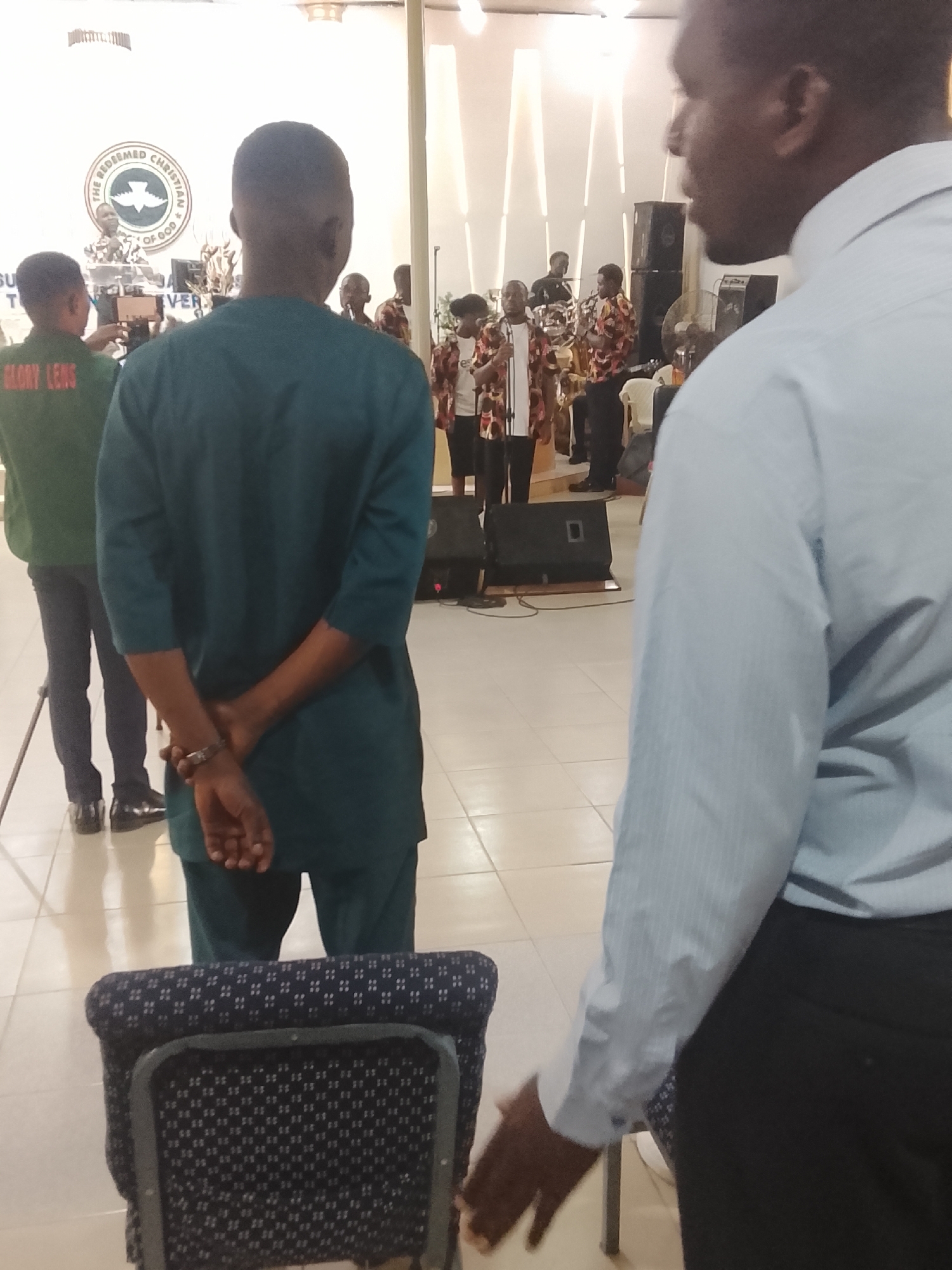 A daytime interior shot shows a church service in progress. The logo of "The Redeemed Christian Church of God" is visible in the background.
Several people are on a raised stage. Some are dressed in matching patterned shirts with black pants, and others are in white tops with black pants. They appear to be part of a choir or musical group.
In the foreground, the backs of two individuals dominate the frame. One, to the left, is wearing a teal green suit. The other, to the right, is wearing a light blue shirt and dark pants. Both face the stage. A folding chair is situated between them.
Another person is visible to the left, wearing a green jacket labeled "GLORY LENS". They are holding a camera on a tripod.
Speakers are placed on the floor in front of the stage. The floor is tiled.
The location is Unknown, Unknown.