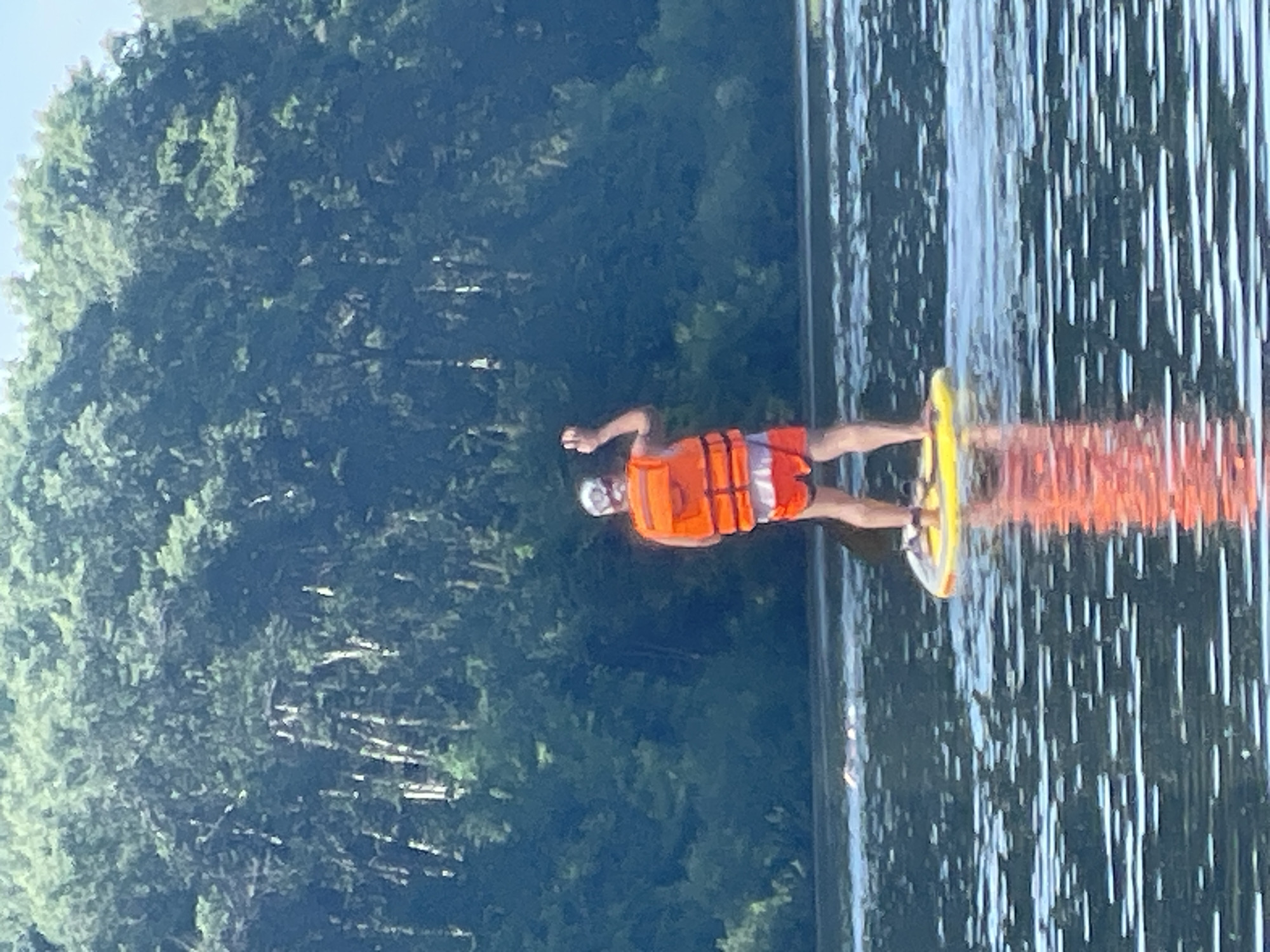 The image shows an individual paddleboarding on a body of water within the city of Vologda, Russia. The person, viewed from behind, wears an orange life vest over red and white striped shorts, and has white hair or a white cap. They stand on a yellow paddleboard, with their right arm raised and hand open, while a black paddle is partially visible in their left hand. Several small, light-colored objects, appearing to be the heads of swimmers, are visible in the mid-distance on the water to the left of the paddleboarder. The background is dominated by a dense wall of dark green deciduous trees, some with lighter trunks, indicating a forested shoreline. The water's surface reflects the bright sky and the orange life vest.