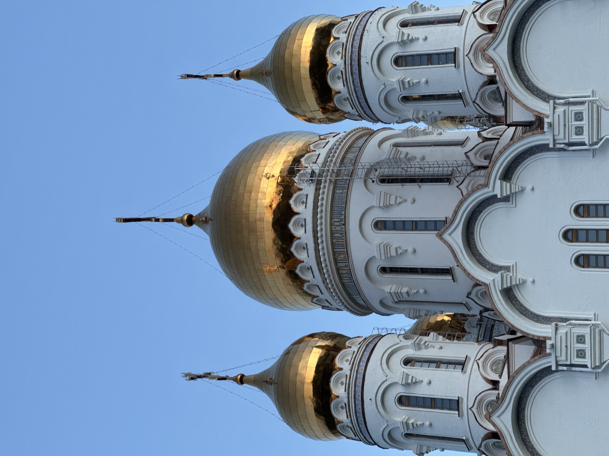 The image shows the domes of the Holy Resurrection Cathedral, located in Starocherkassk, Russia. The domes are golden and reflect sunlight. The cathedral is white and features architectural details such as arches, windows, and decorative elements. The background is a clear blue sky. The image appears to be taken from a low angle, emphasizing the height of the cathedral.