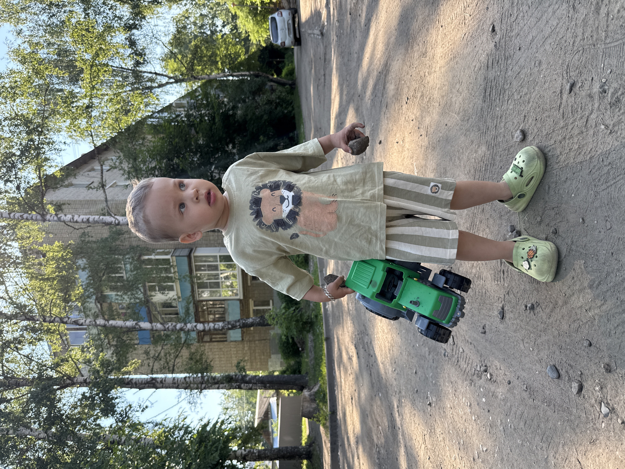 A young Caucasian boy, appearing to be around 2-3 years old, stands on a gravelly path in front of a multi-story building with a light-colored brick facade and blue trim. The boy is wearing a light green t-shirt with a cartoon lion graphic and light green and white striped shorts. He has on light green clogs with decorative charms. In his right hand, he holds a small, dark-colored rock, and in his left arm, he carries a bright green toy tractor. The setting suggests an outdoor urban environment, possibly a residential courtyard, with trees and shadows cast by the sun. A white car is visible in the background on a paved road. The boy is looking to his right, with a curious expression. The overall scene is illuminated by natural sunlight.
