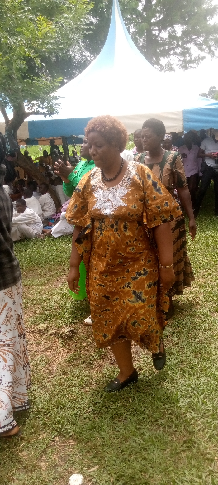 The image shows a gathering of people outdoors, likely at a celebration or event. The main subject is a woman wearing a brown and black patterned dress with white embellishments. She stands on a patch of green grass, with other people behind her, under a large white tent with blue accents. A few other people are visible in the background, with some sitting and others standing. The woman is walking toward the right side of the image with her left foot slightly raised, showing a black leather loafer with a decorative detail.
The location of the scene is within the city of Burundu, Kenya, based on the provided coordinates (0.459, 34.776). The event is held in a grassy outdoor area, likely a park or a public space. The overall mood appears celebratory, although the individual’s facial expression is unclear. The focus is on the woman in the brown and black dress. The image was likely taken during daylight hours.